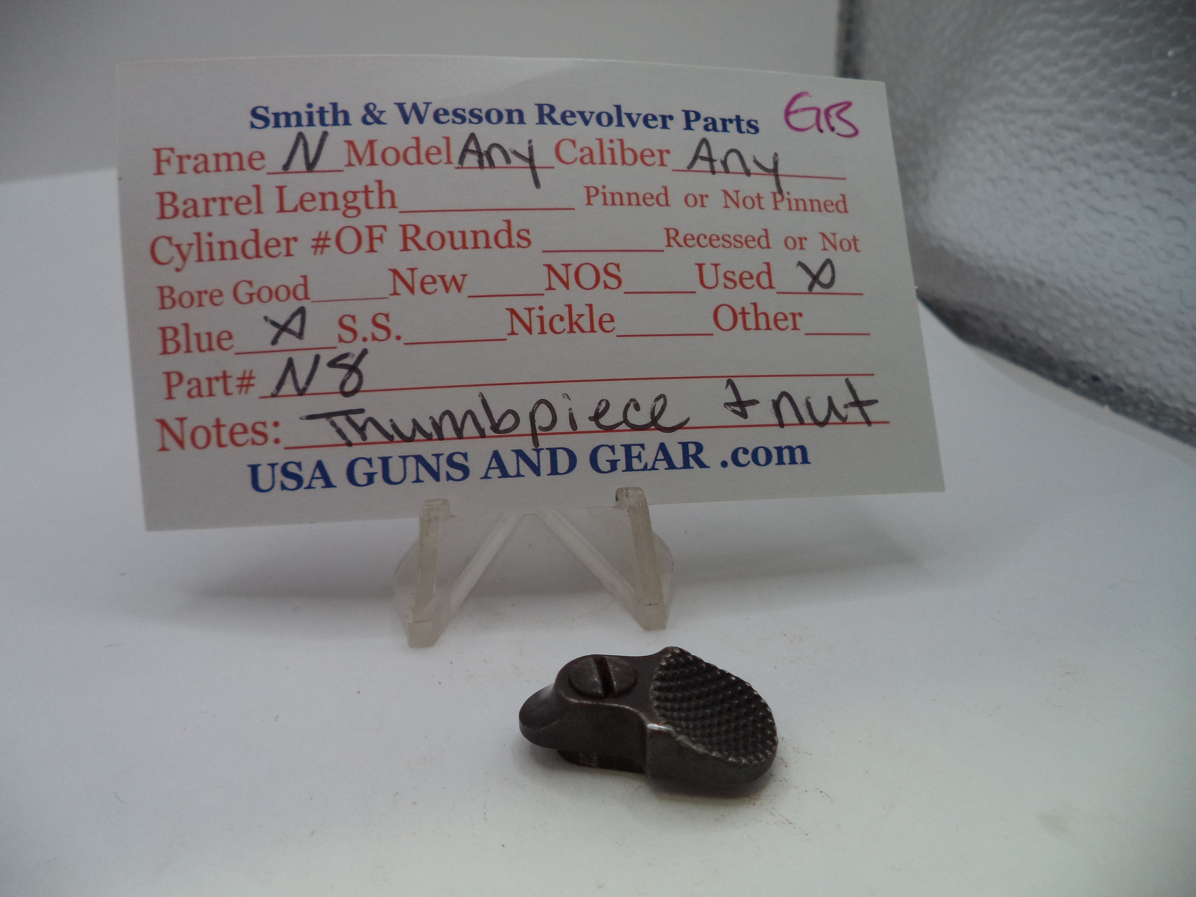 A small dark metal revolver part displayed on a clear stand in front of a printed parts information card for Smith & Wesson revolvers, with handwritten notes on the card.