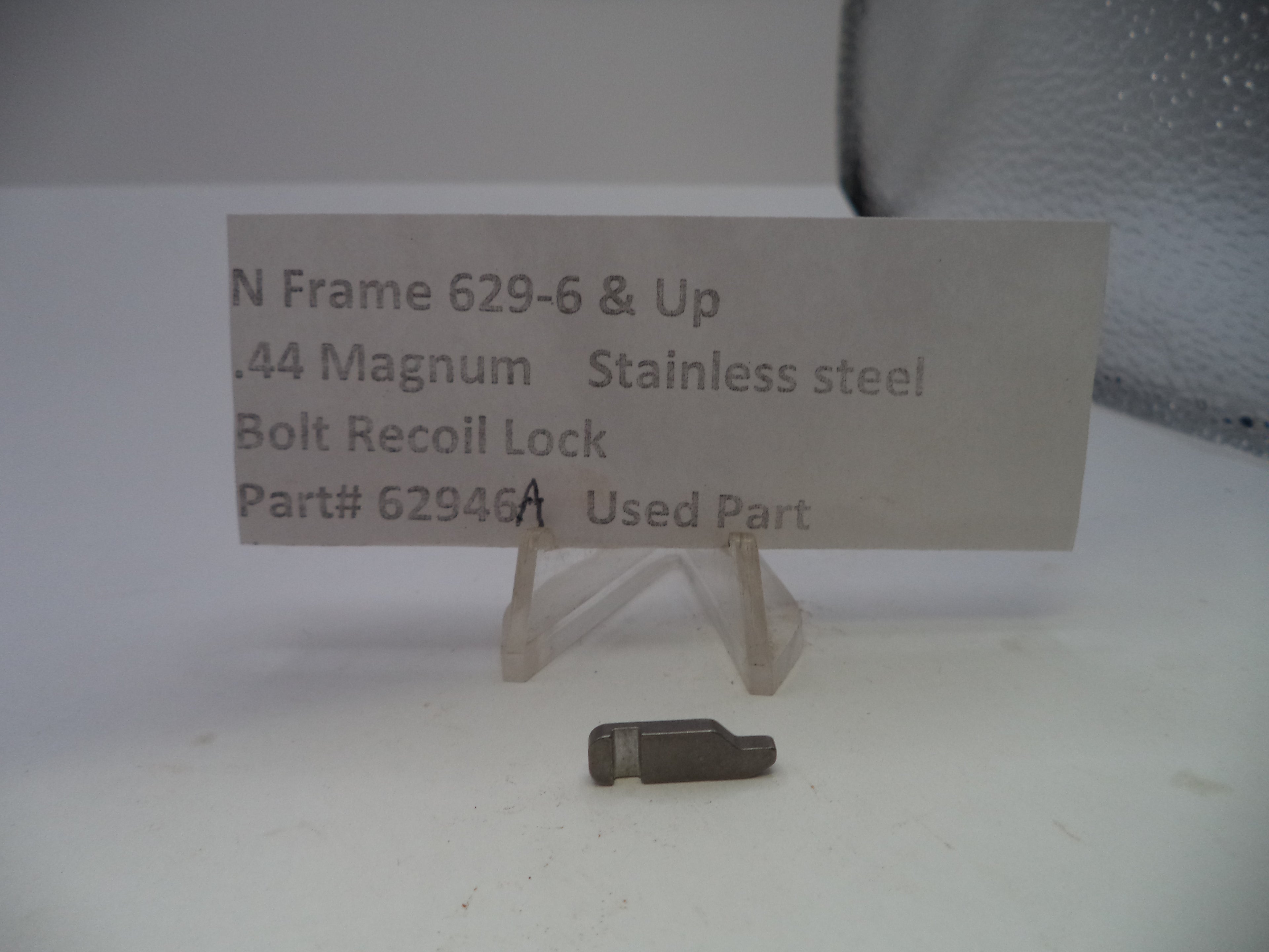 A small used stainless steel bolt recoil lock for a .44 Magnum rifle, displayed on a clear plastic stand next to a paper label card with part details.