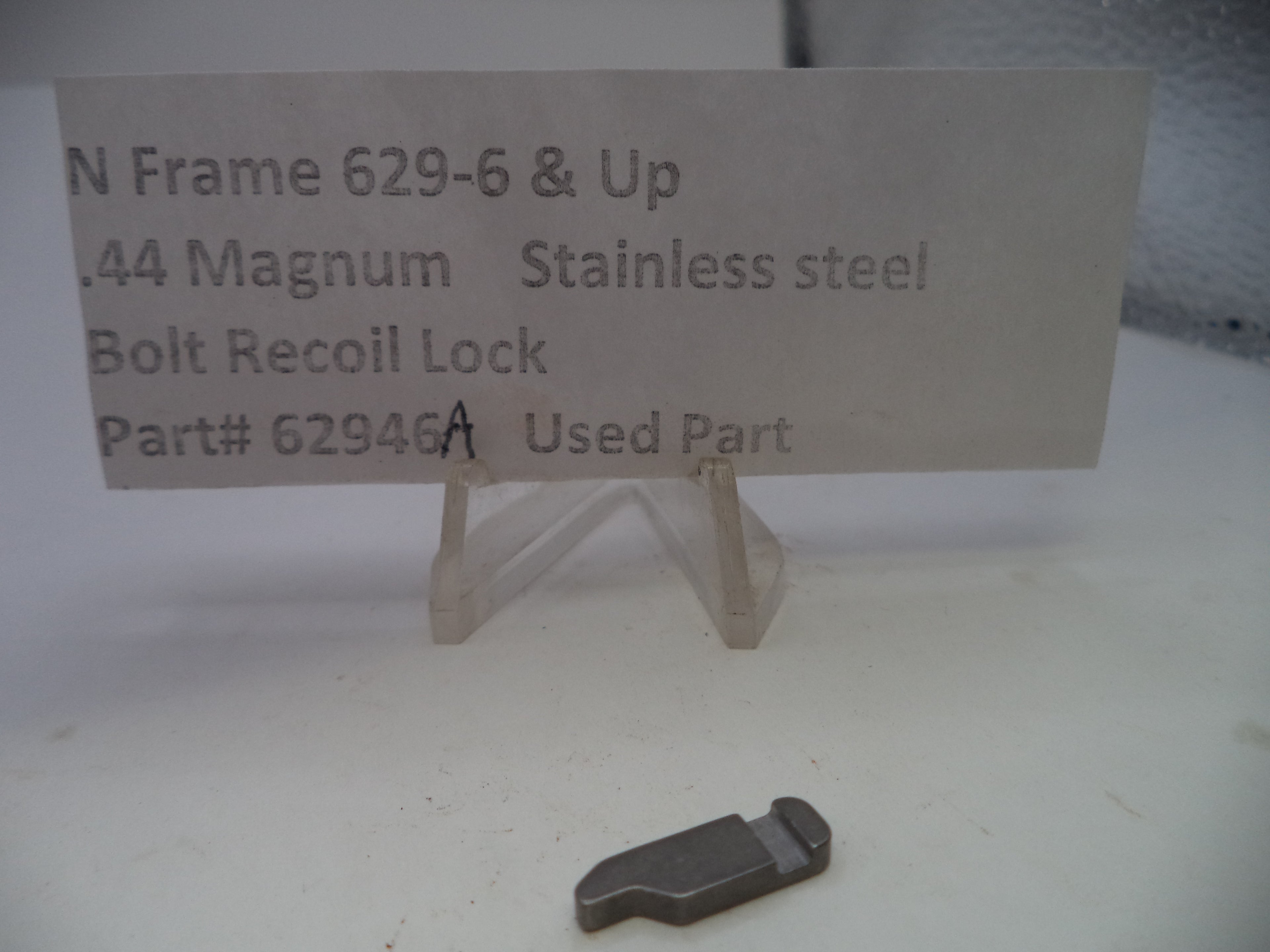 A used stainless steel bolt recoil lock for a .44 Magnum N Frame, displayed on a small clear stand next to a paper label card with part number 62946A.