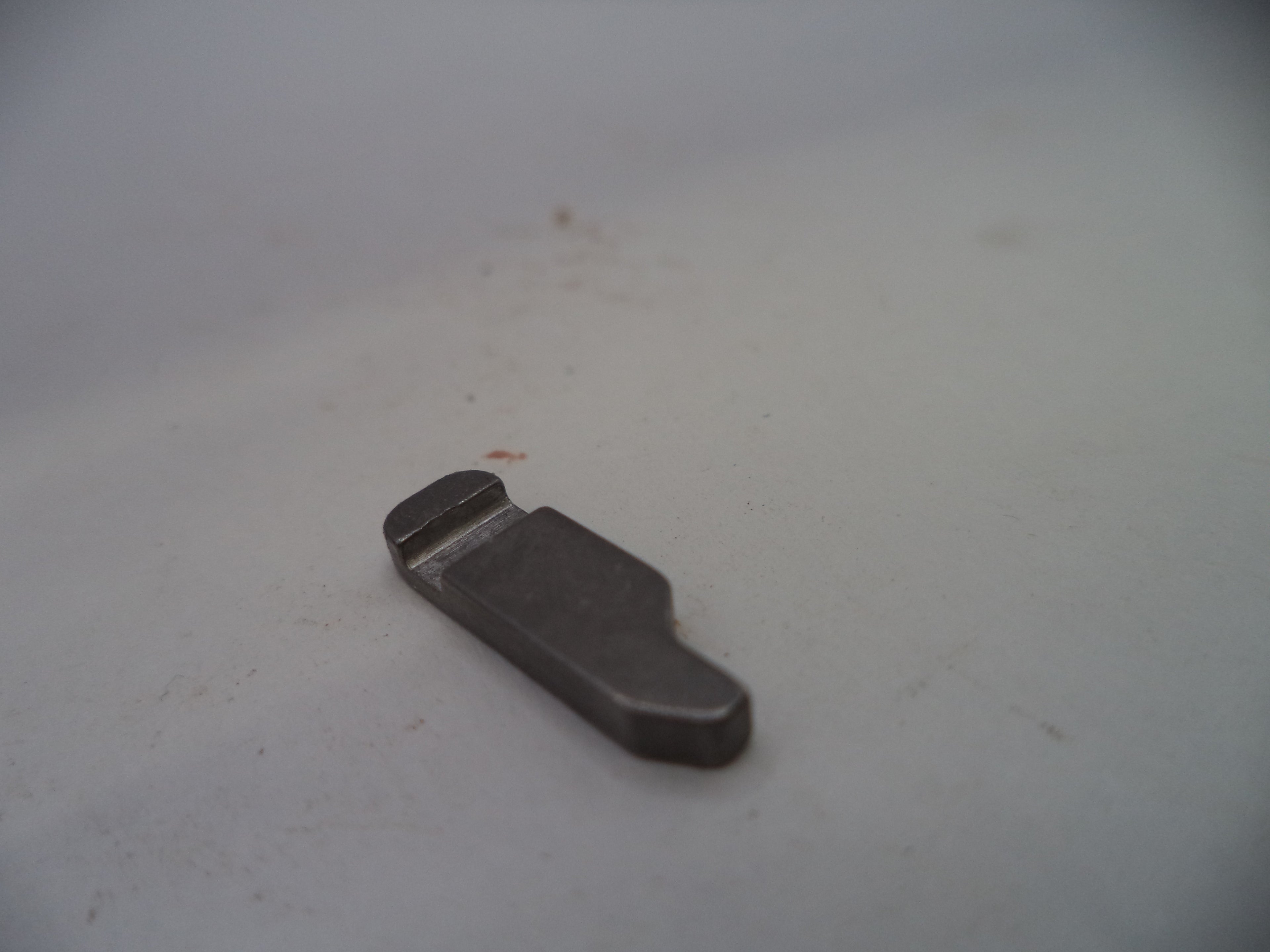 A small dark gray metal replacement part, possibly a clip or bracket, resting on a light gray surface.