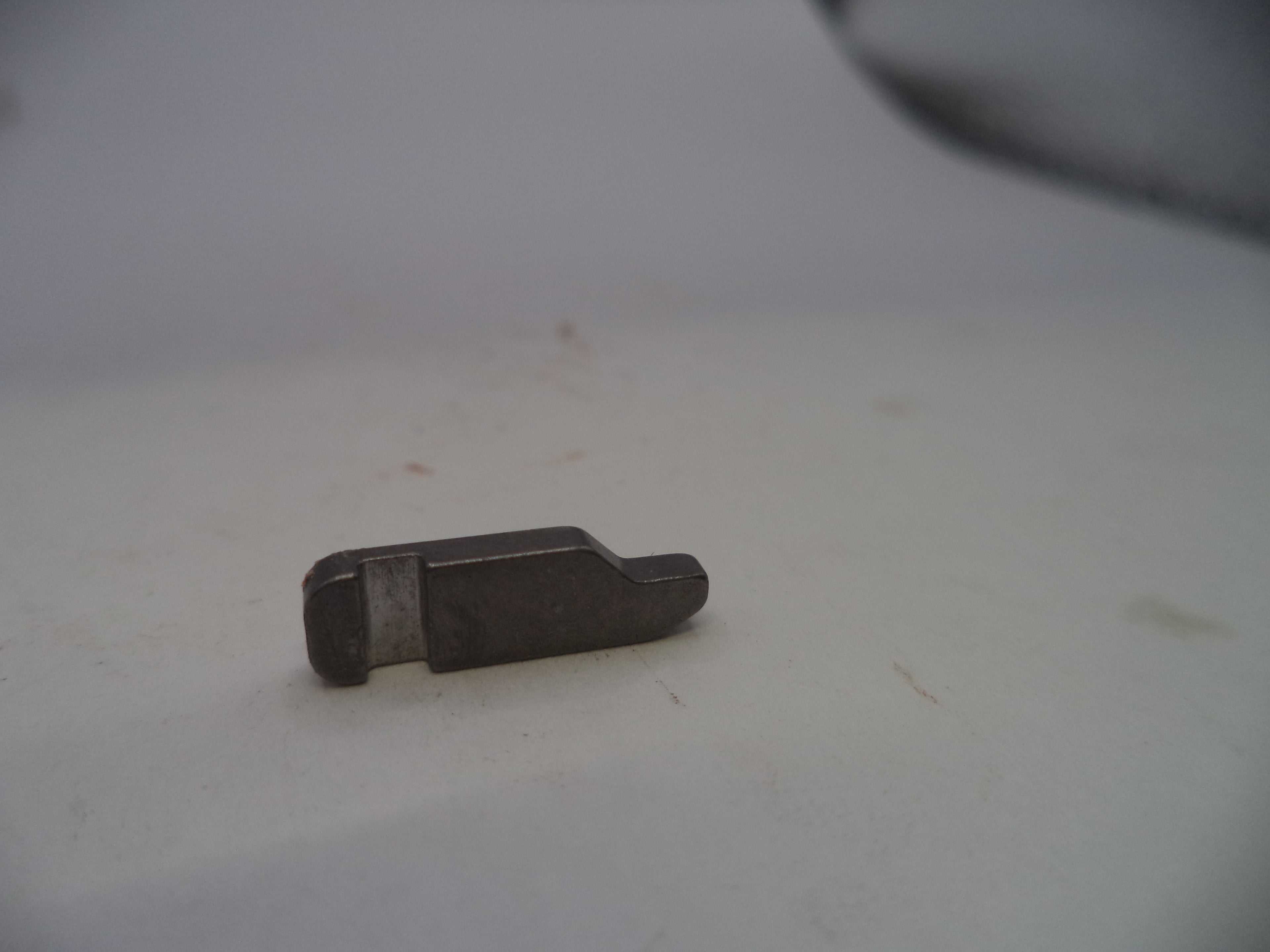 A small, dark metallic replacement part, possibly a tool bit or mechanical component, resting on a light gray surface.