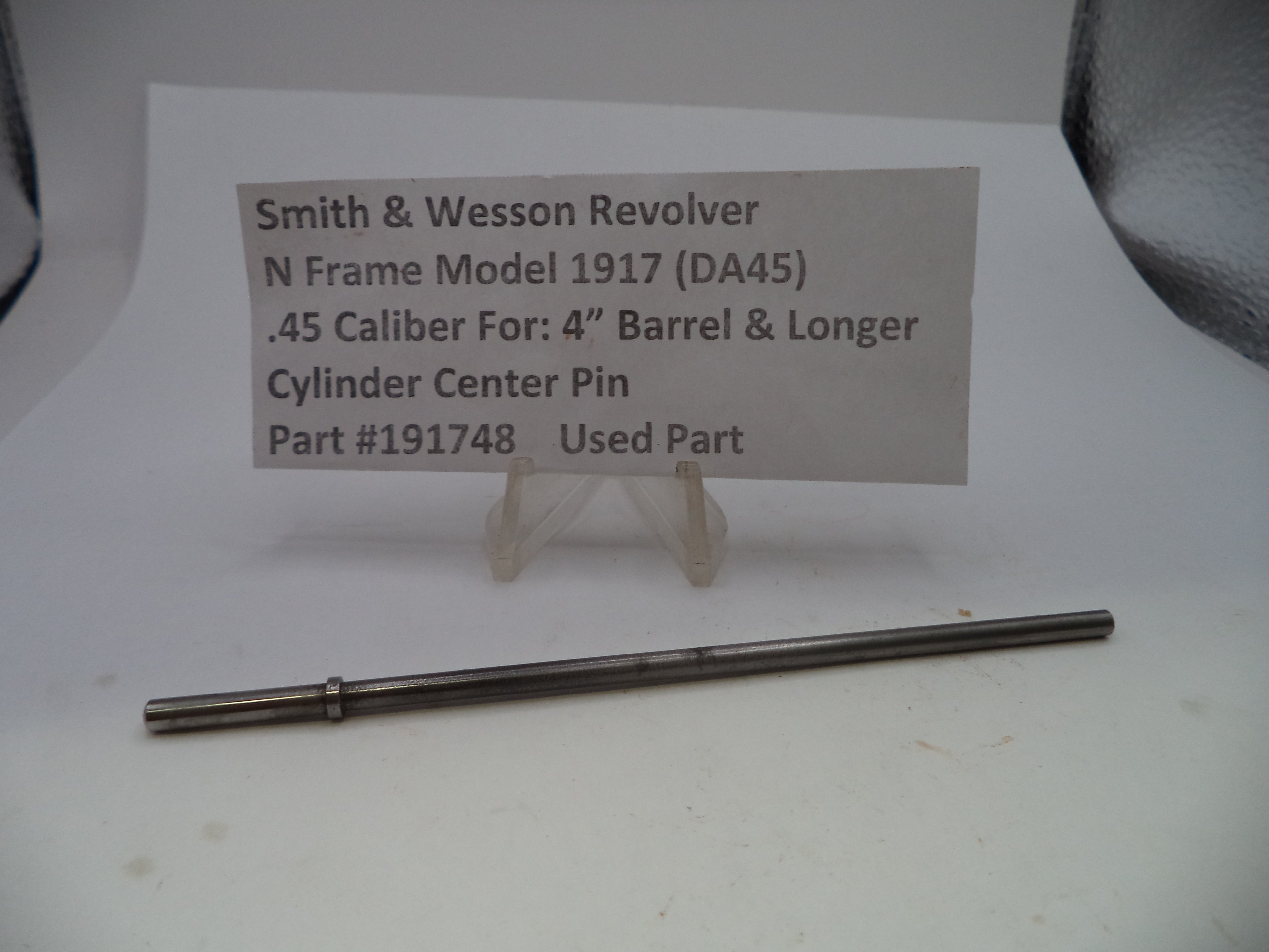 A used metal cylinder center pin for a Smith & Wesson revolver, displayed on a white surface below a paper label card with part details.