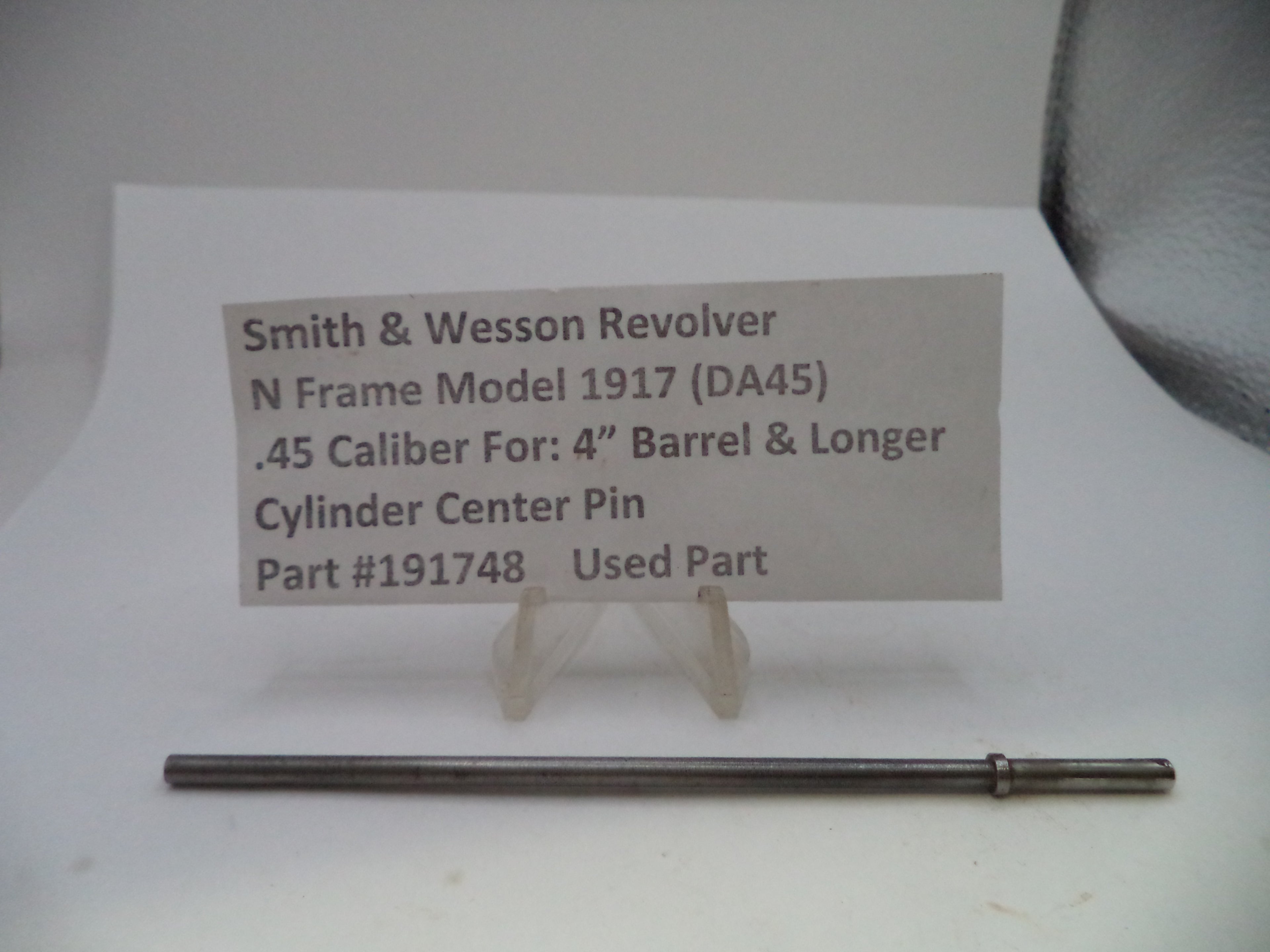 A used metal cylinder center pin for a Smith & Wesson revolver, displayed on a white surface below a paper label card with part details.