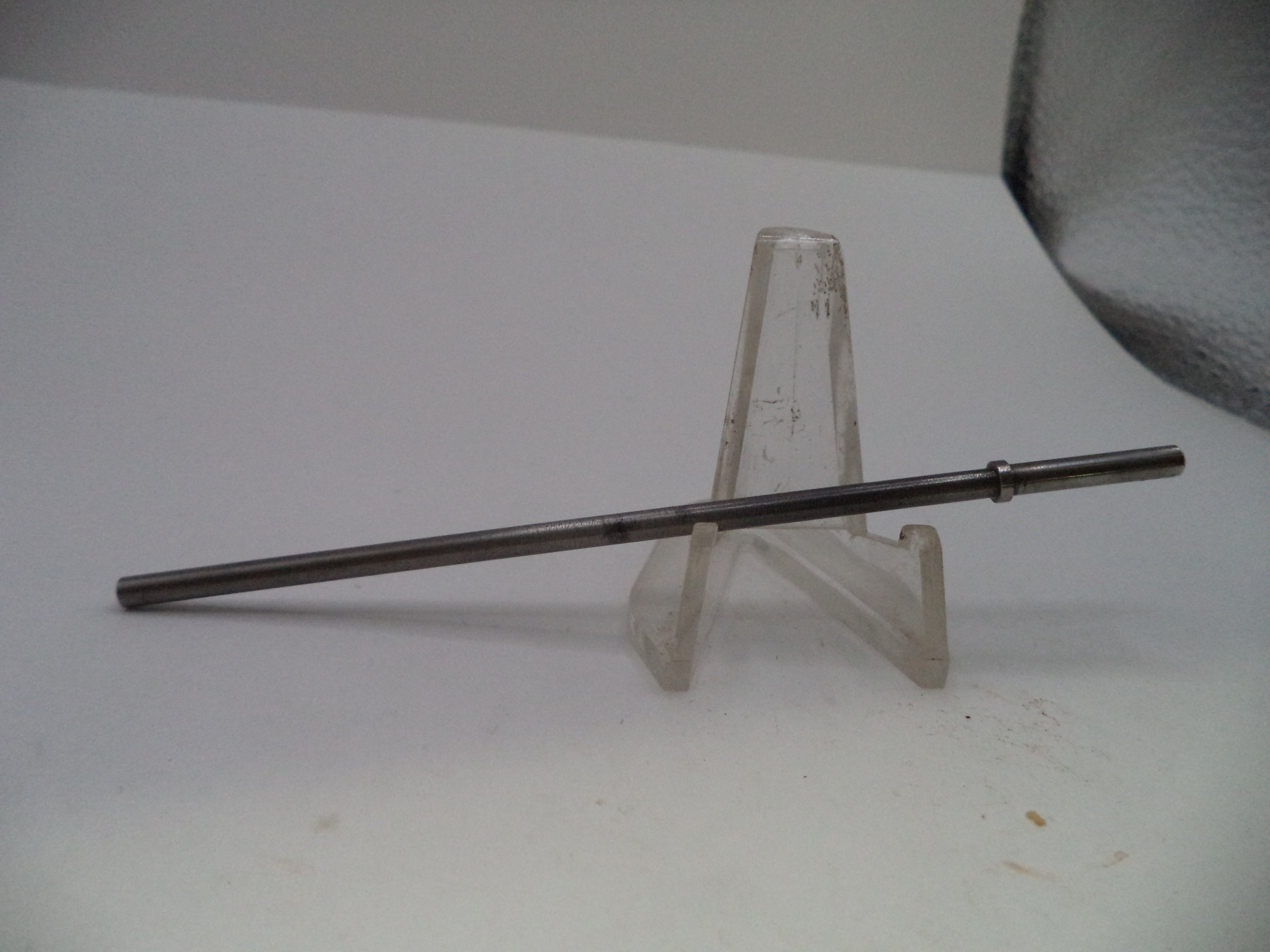 A long, thin metallic rod or pin displayed horizontally on a clear plastic stand against a plain white background.