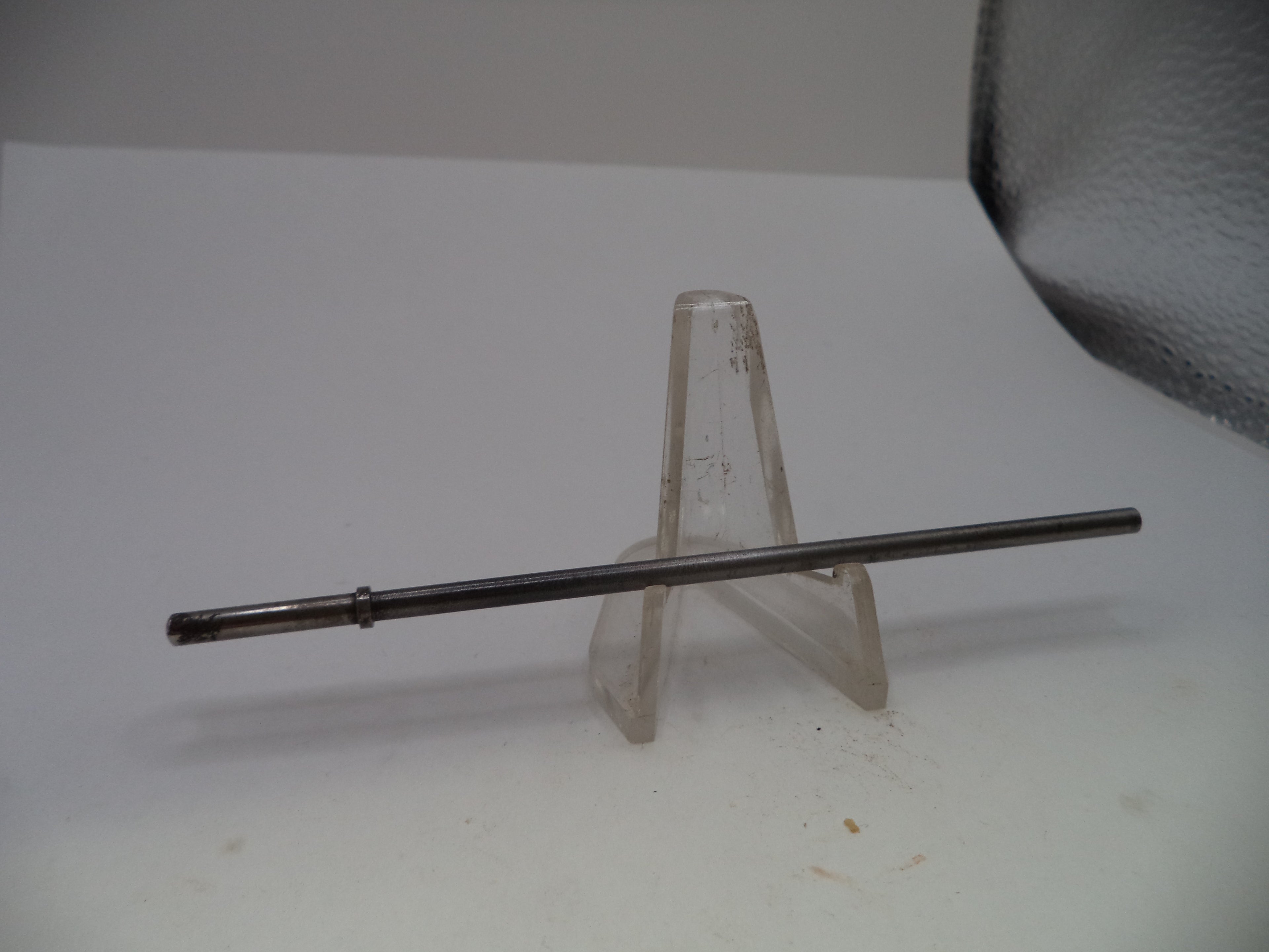 A long, thin dark metallic rod or pin displayed horizontally on a clear plastic stand against a plain light background.