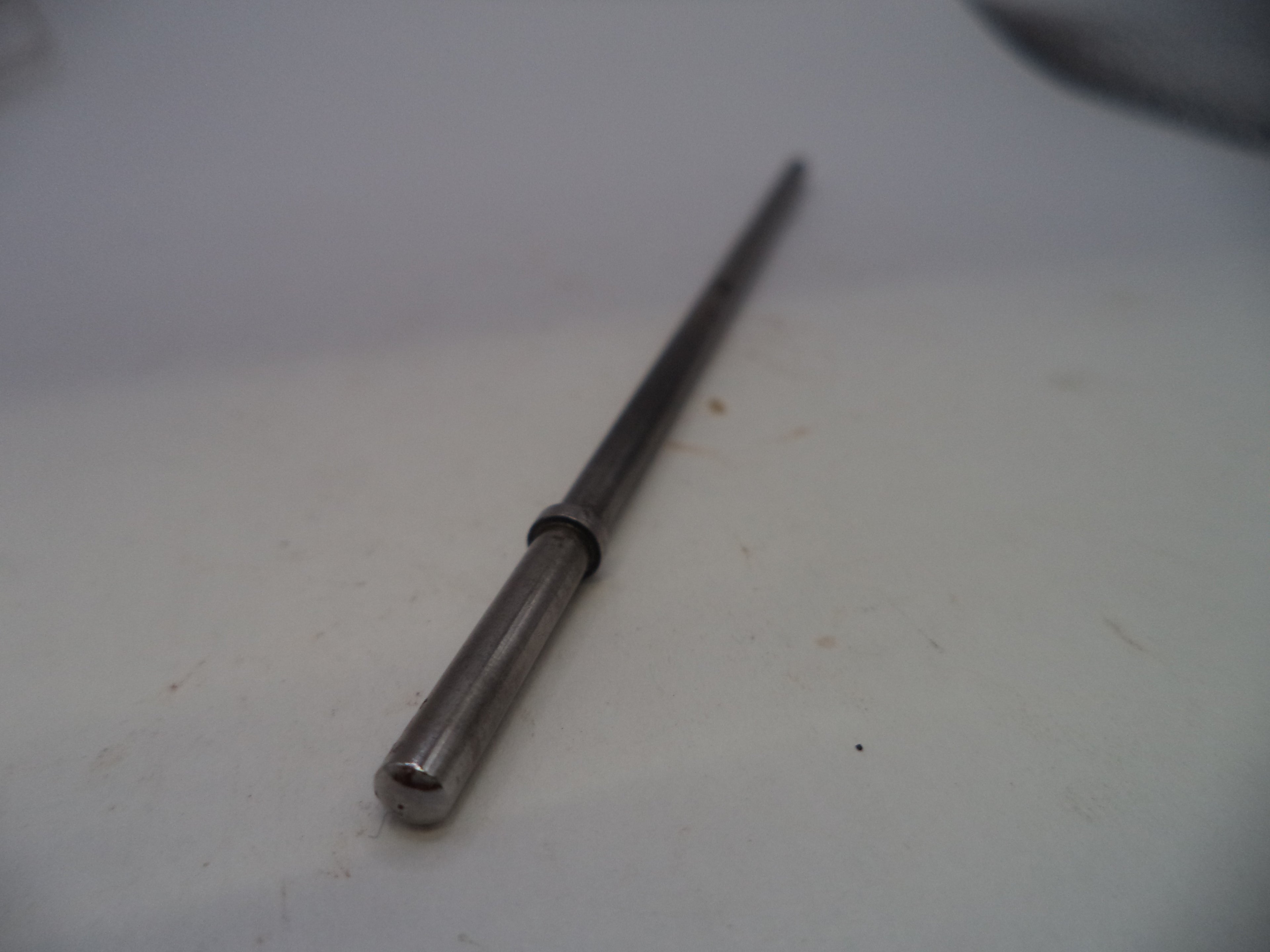A long, slender metallic rod or shaft with a cylindrical end and a small dark ring near the center, photographed on a plain light surface.
