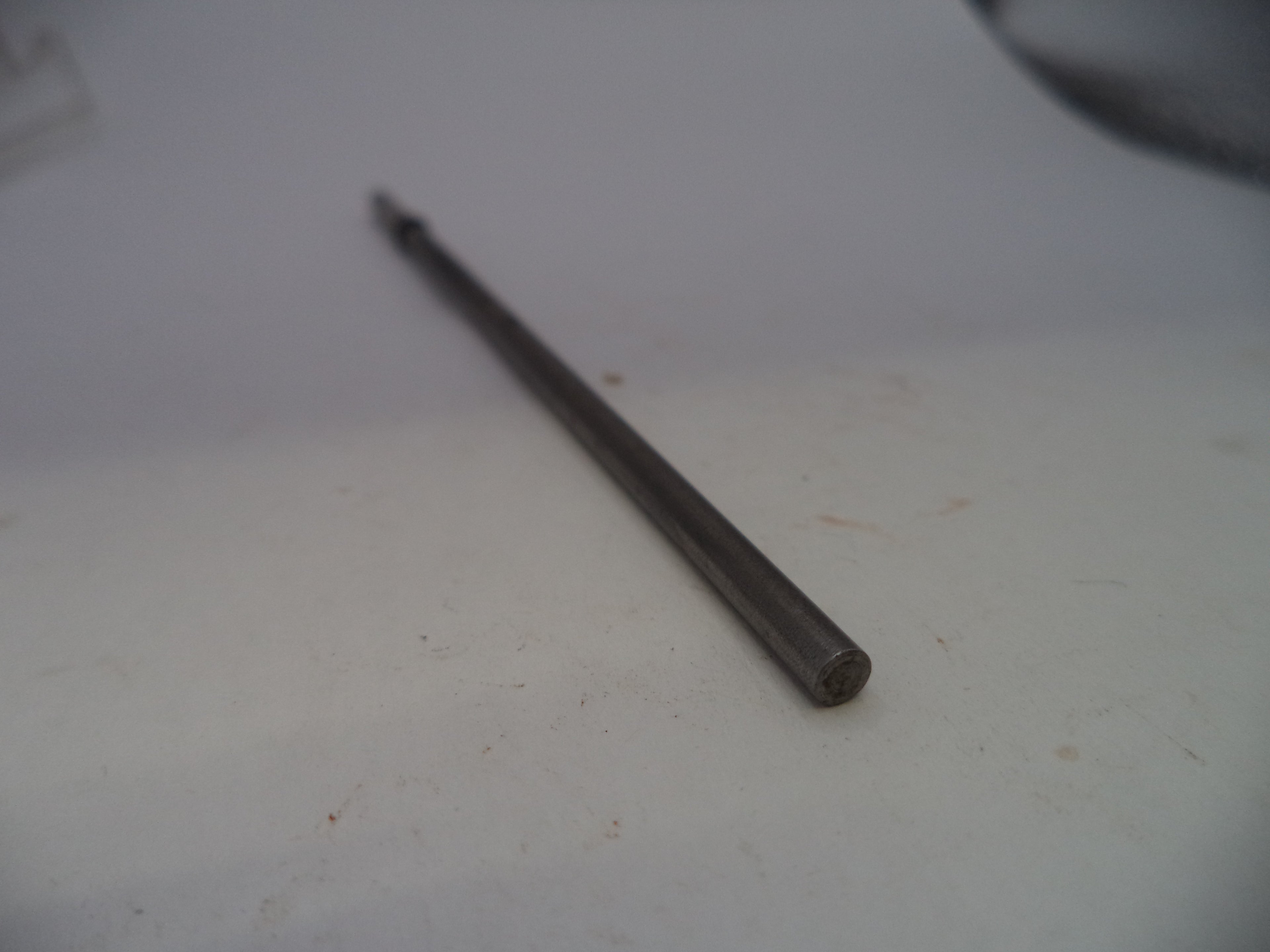 A single dark metallic screwdriver bit or small tool bit placed diagonally on a plain off-white surface.