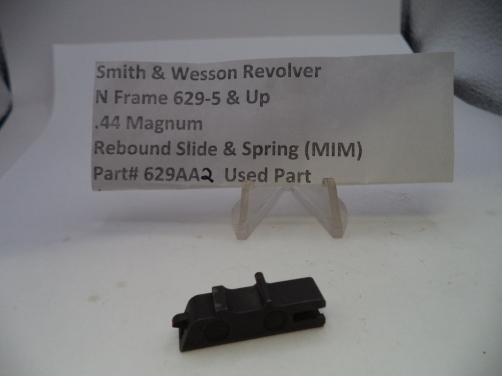 A small black metal revolver rebound slide and spring replacement part displayed on a white surface in front of a clear stand holding a paper label that reads 'Smith & Wesson Revolver N Frame 629-5 & Up .44 Magnum Rebound Slide & Spring (MIM) Part# 629AAQ Used Part'.