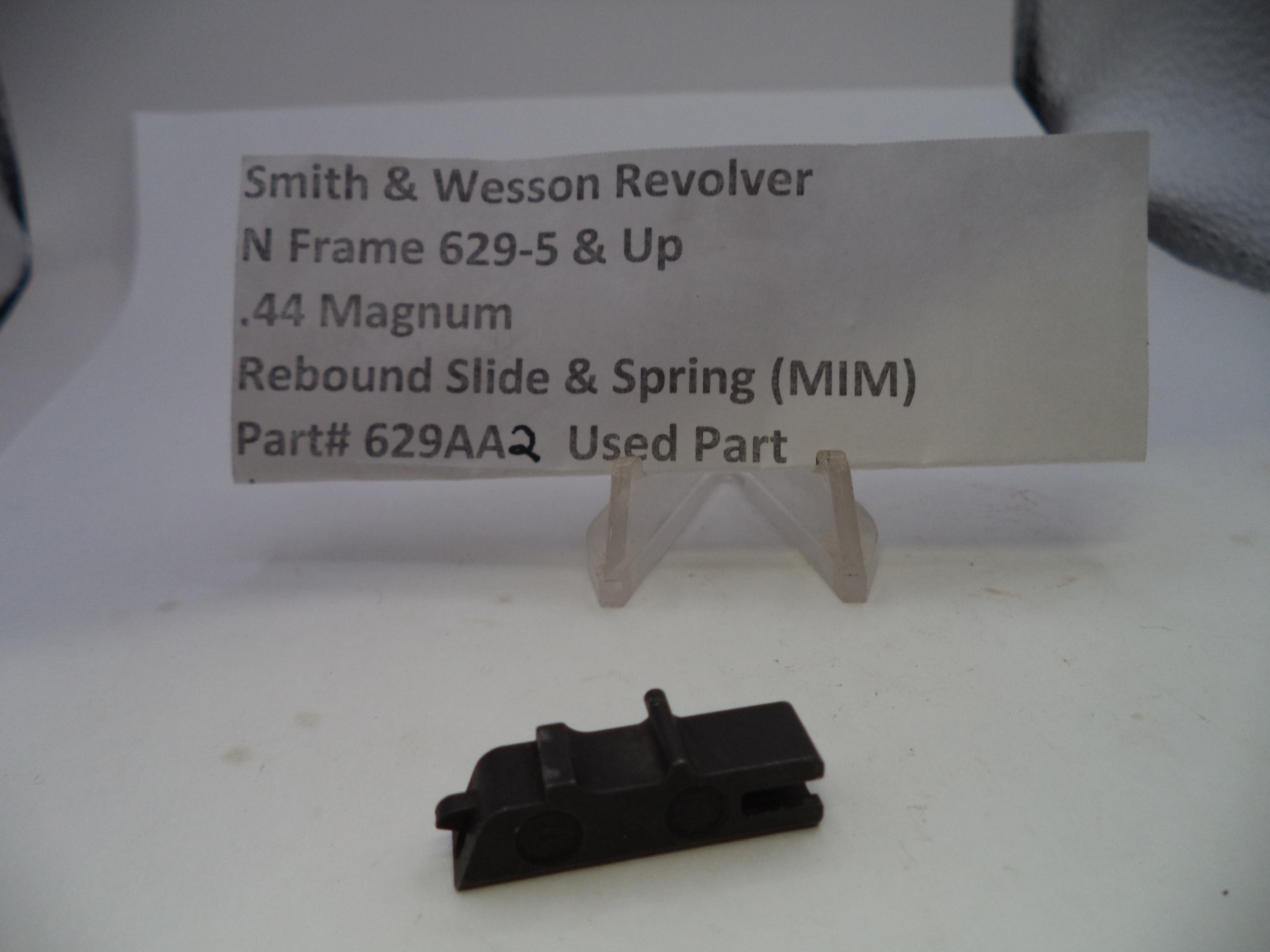 A small black metal revolver rebound slide and spring replacement part displayed on a white surface in front of a clear stand holding a paper label that reads 'Smith & Wesson Revolver N Frame 629-5 & Up .44 Magnum Rebound Slide & Spring (MIM) Part# 629AAQ Used Part'.