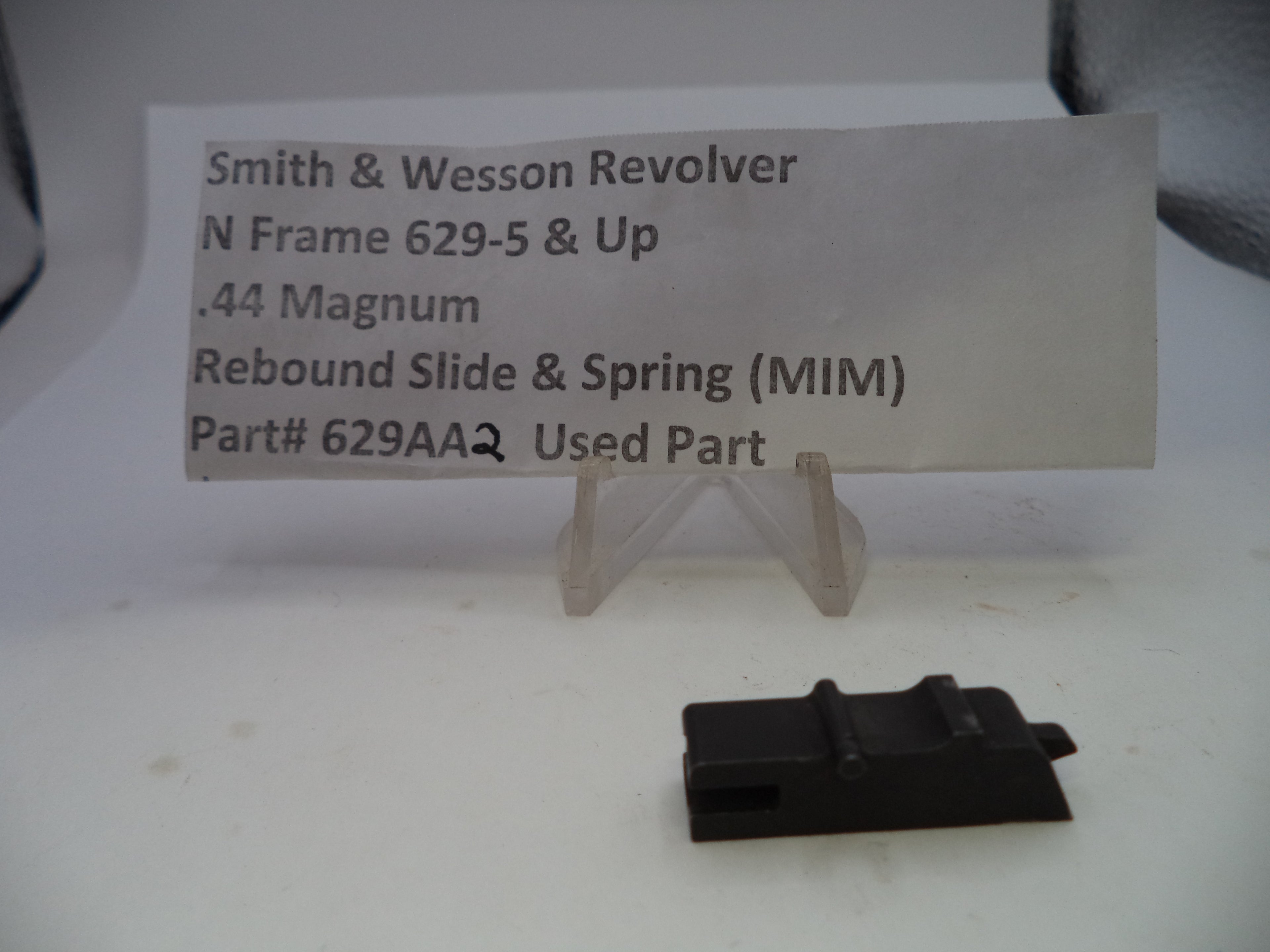 A used black metal Smith & Wesson revolver rebound slide and spring part, displayed on a white surface with a clear plastic stand holding a paper label.