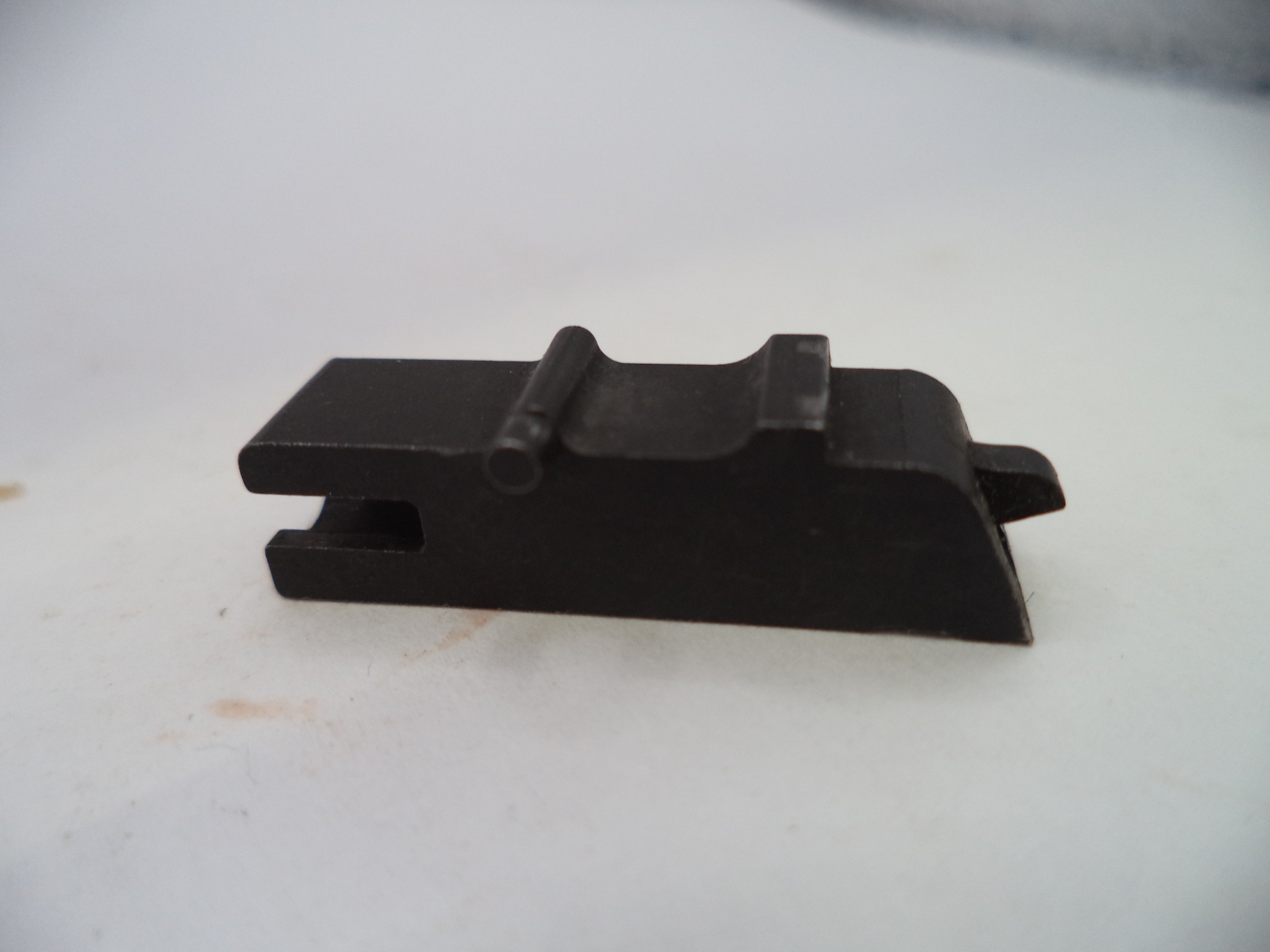 A small black metal component, likely a firearm magazine or accessory part, photographed on a plain off-white surface.