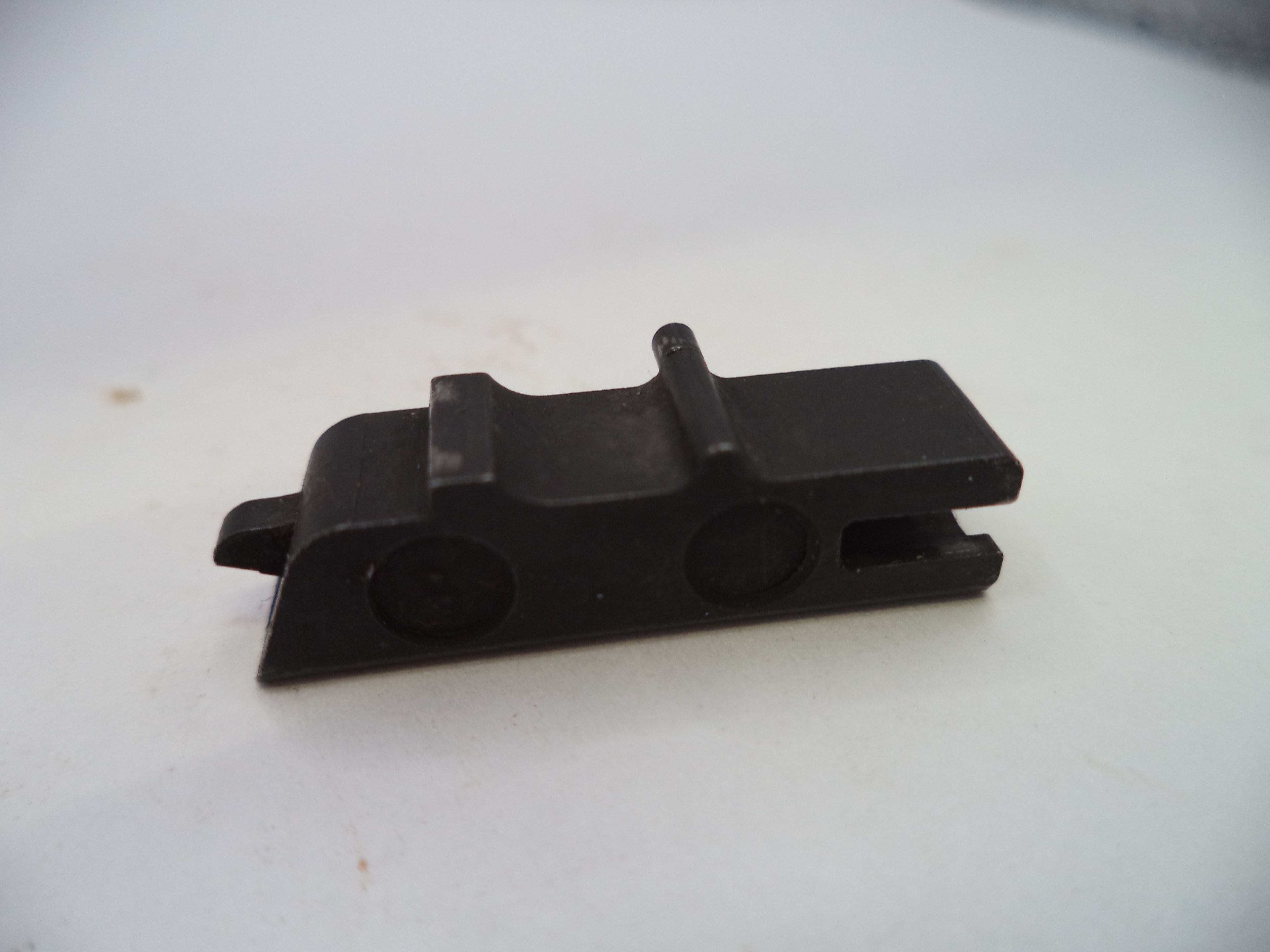 A small black plastic or metal hardware component with a rectangular body, two circular recesses, and a small protruding tab, photographed on a plain light background.