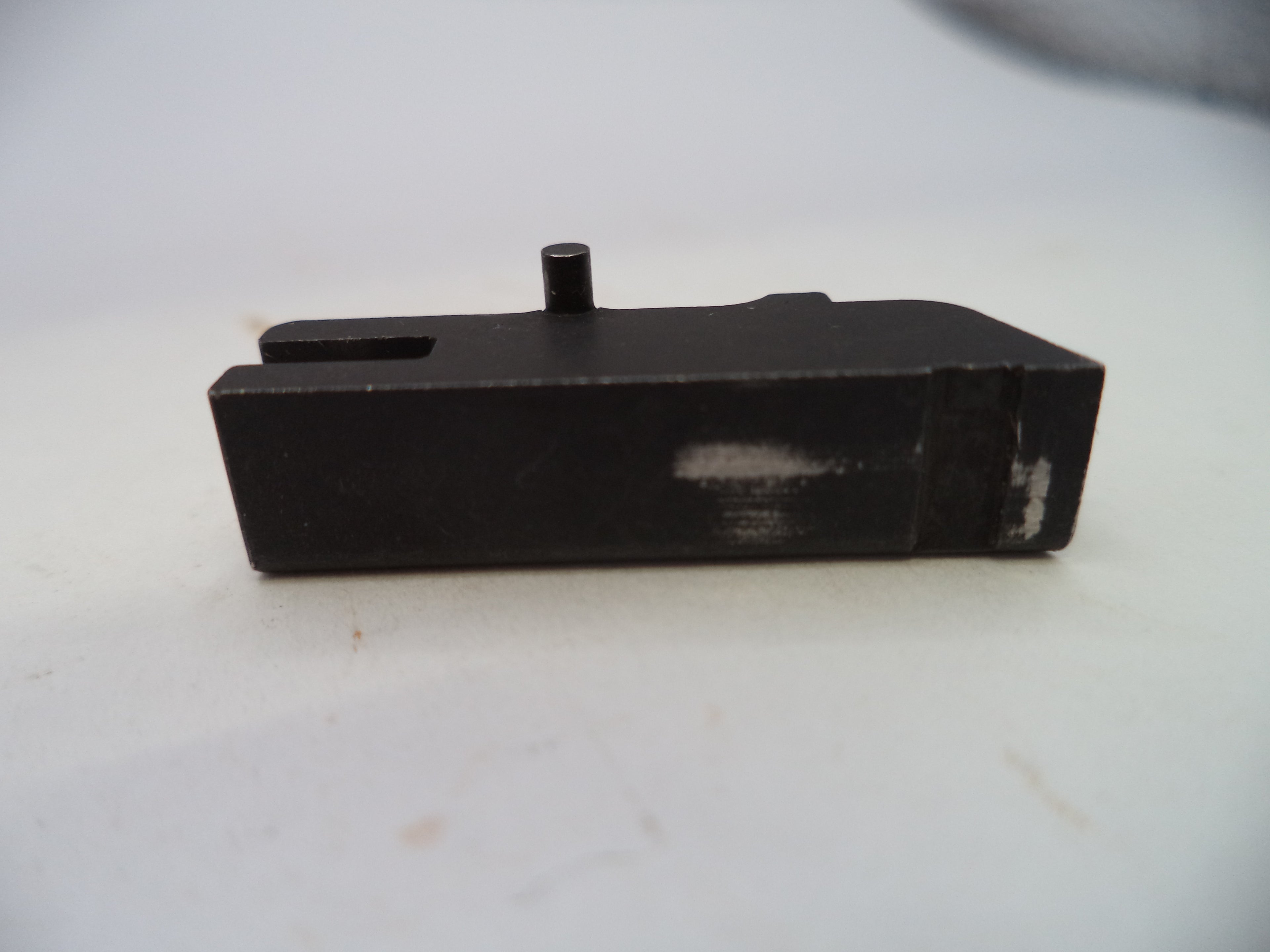 A black metal mechanical component, likely a receiver or magazine body, with a small protruding pin and a rectangular slot, shown on a light gray background.