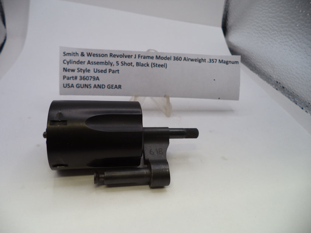 A used black metal cylinder assembly for a Smith & Wesson revolver, displayed on a white surface with a white paper label card behind it.