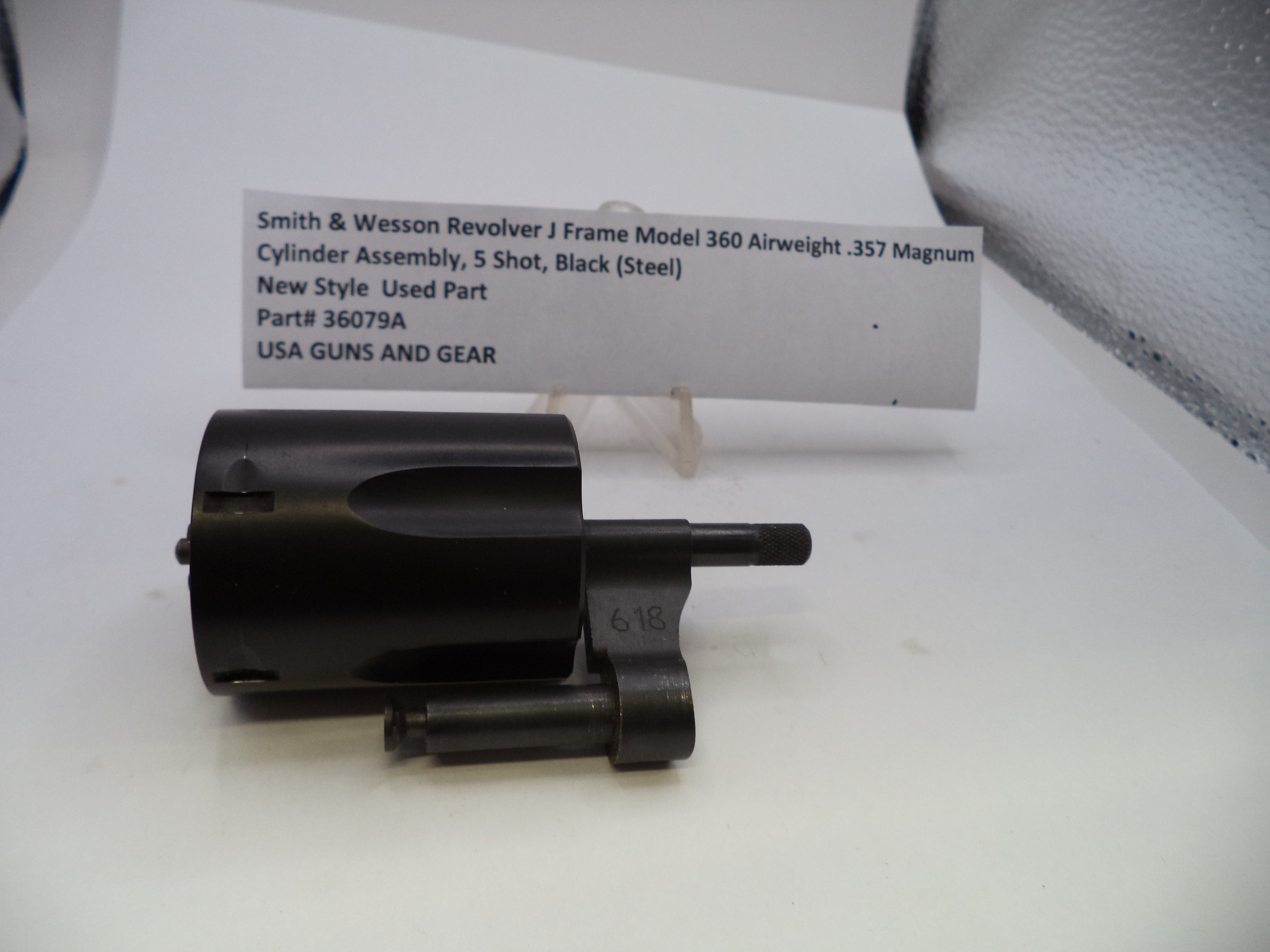 A used black metal cylinder assembly for a Smith & Wesson revolver, displayed on a white surface with a white paper label card behind it.