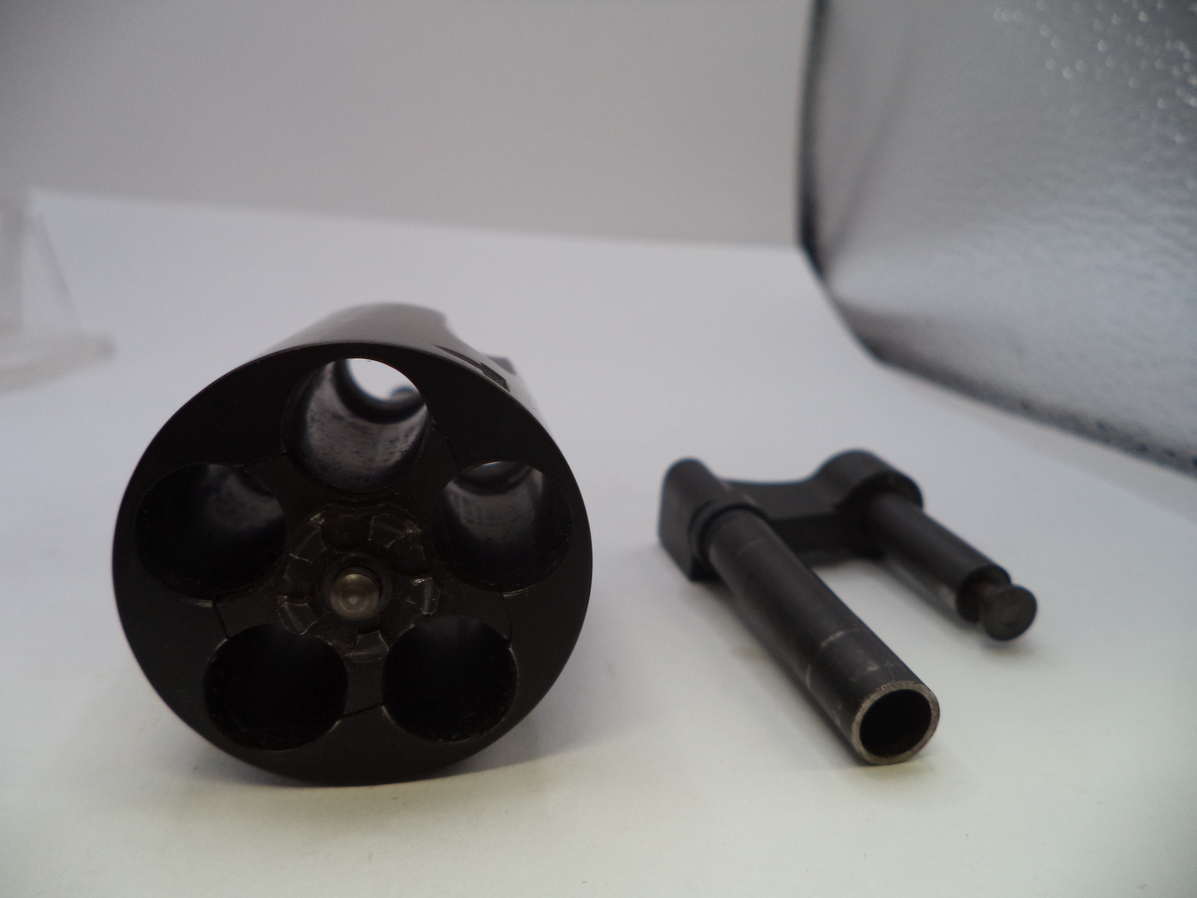 A close-up of a black metal revolver cylinder disassembled to show the internal barrel holes, with a small black metal tool placed beside it on a white surface.