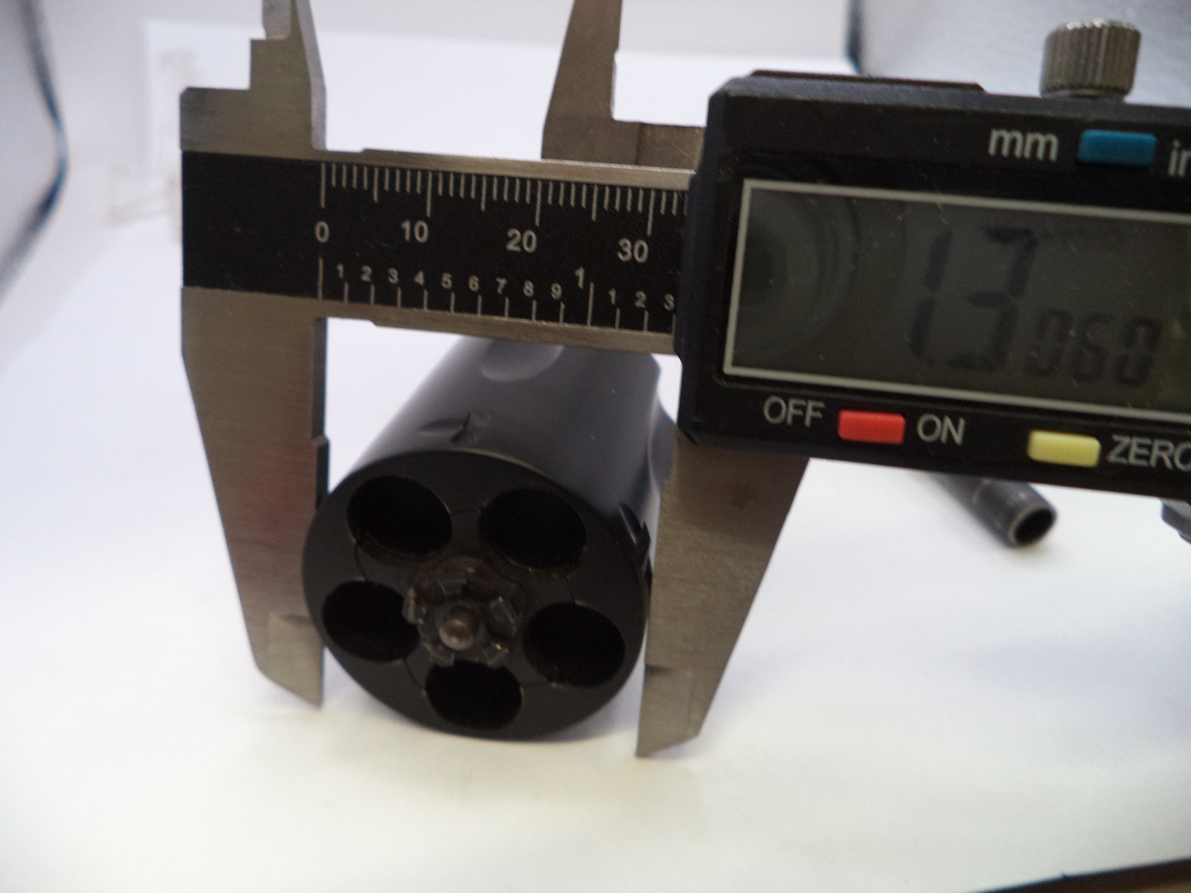 A close-up of a black cylindrical component with five circular openings, measured with a digital caliper showing 13.960 mm on a white background.