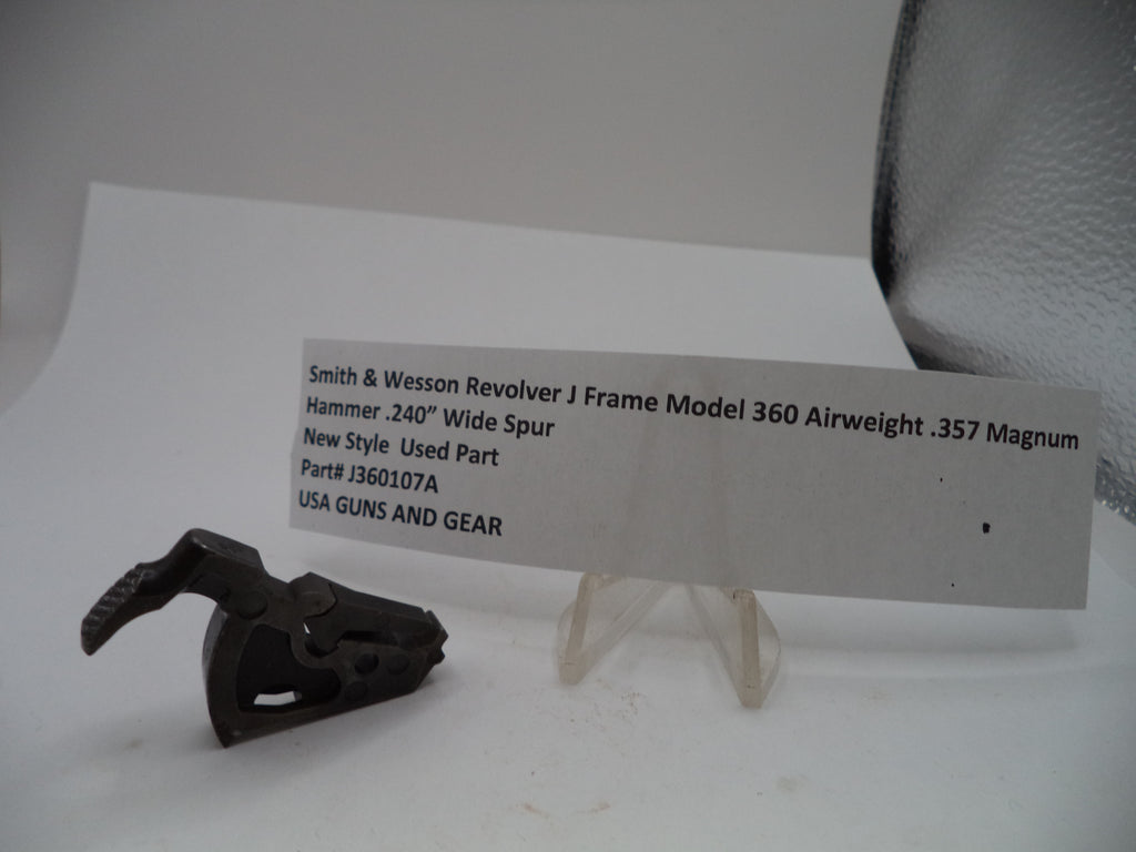 A used black metal revolver hammer part displayed next to a white paper label card on a clear stand against a plain light background.