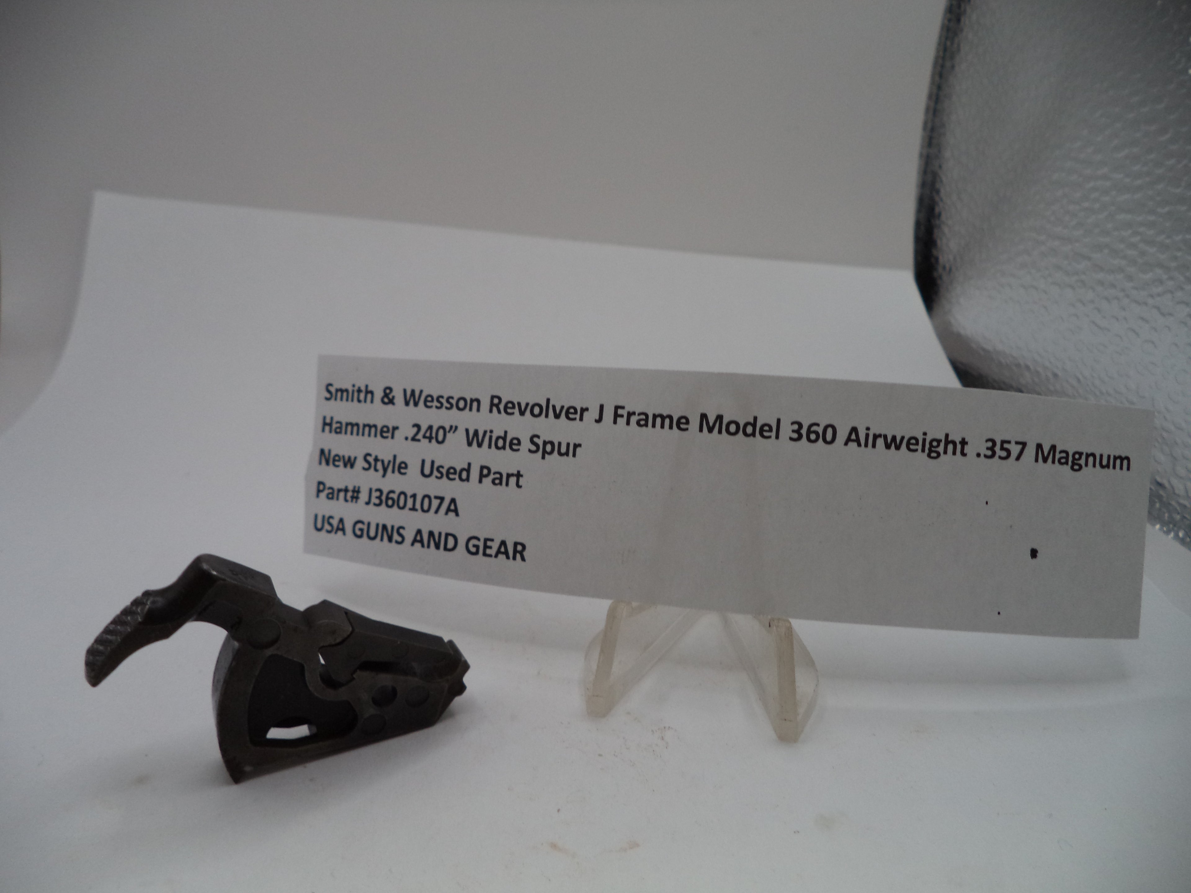 A used black metal revolver hammer part displayed next to a white paper label card on a clear stand against a plain light background.