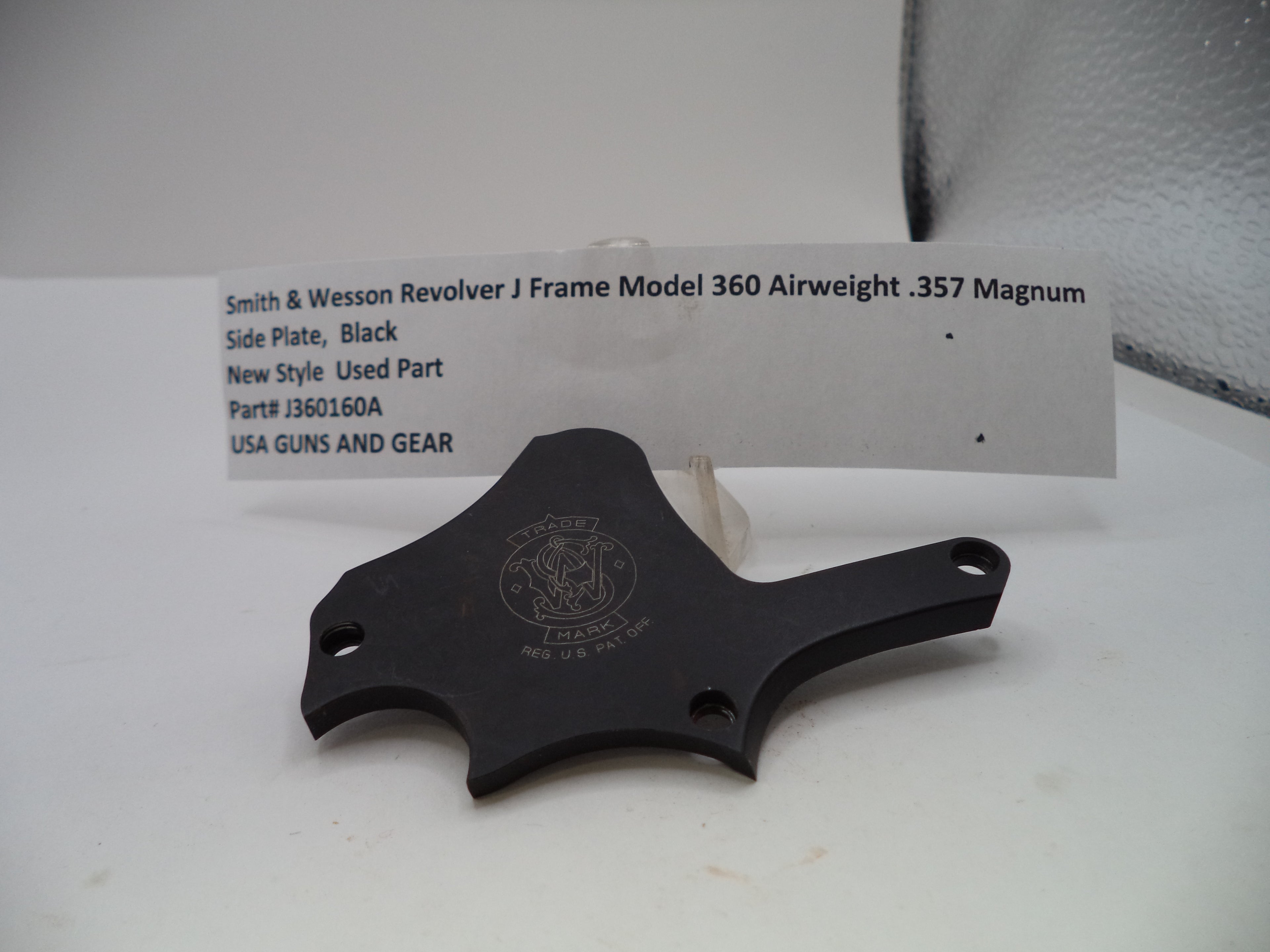 A black metal Smith & Wesson revolver J frame side plate part, displayed on a white surface with a white product information card behind it.
