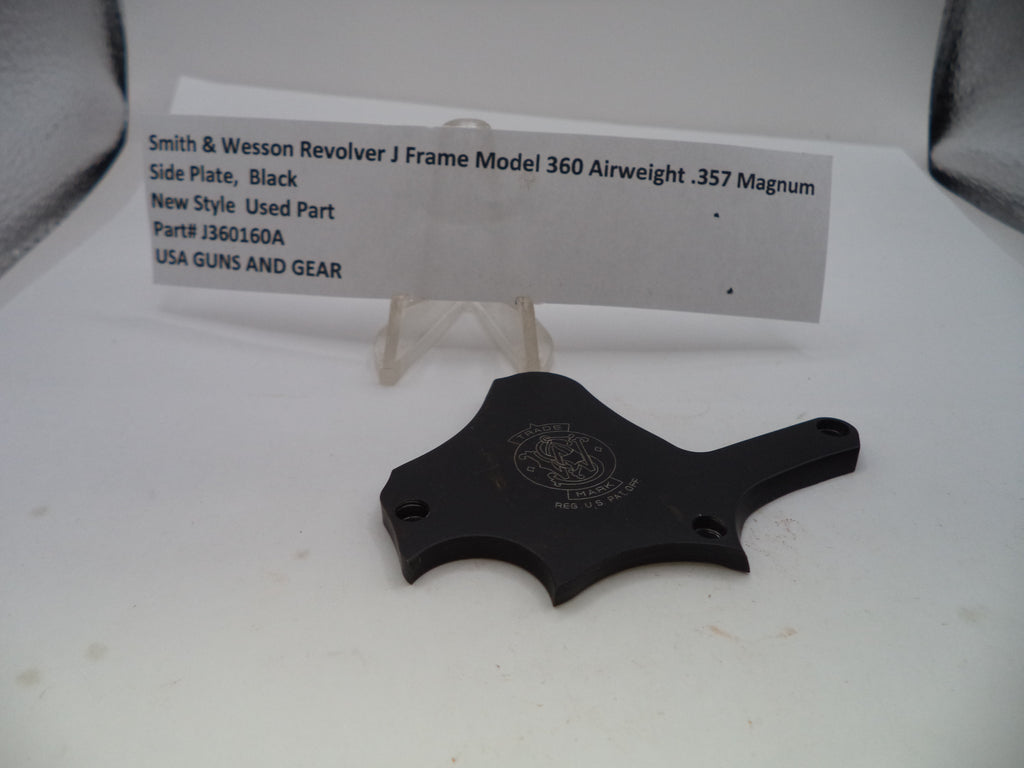 A black metal side plate for a Smith & Wesson revolver, displayed on a white surface with a small white label card behind it.