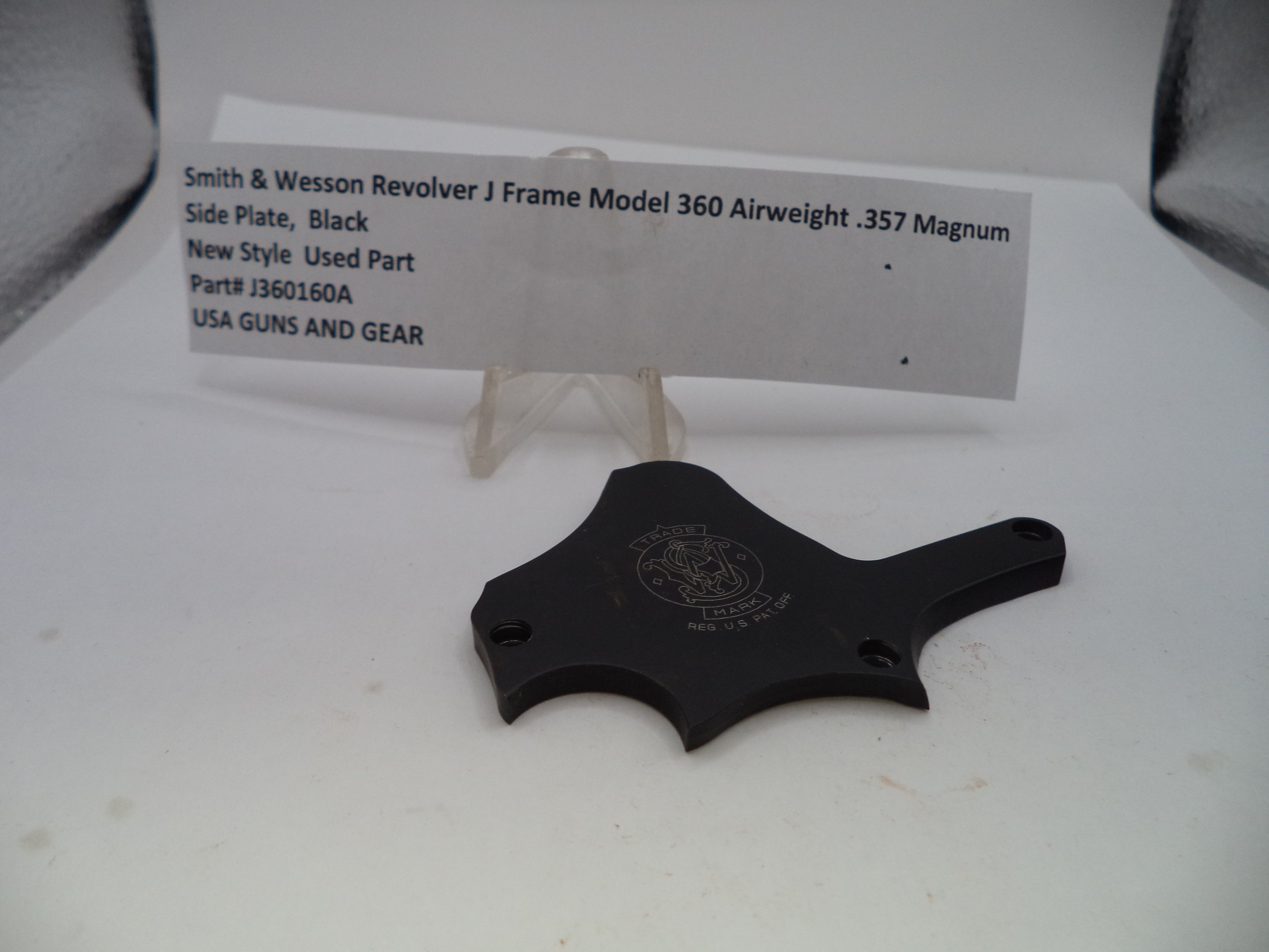 A black metal side plate for a Smith & Wesson revolver, displayed on a white surface with a small white label card behind it.