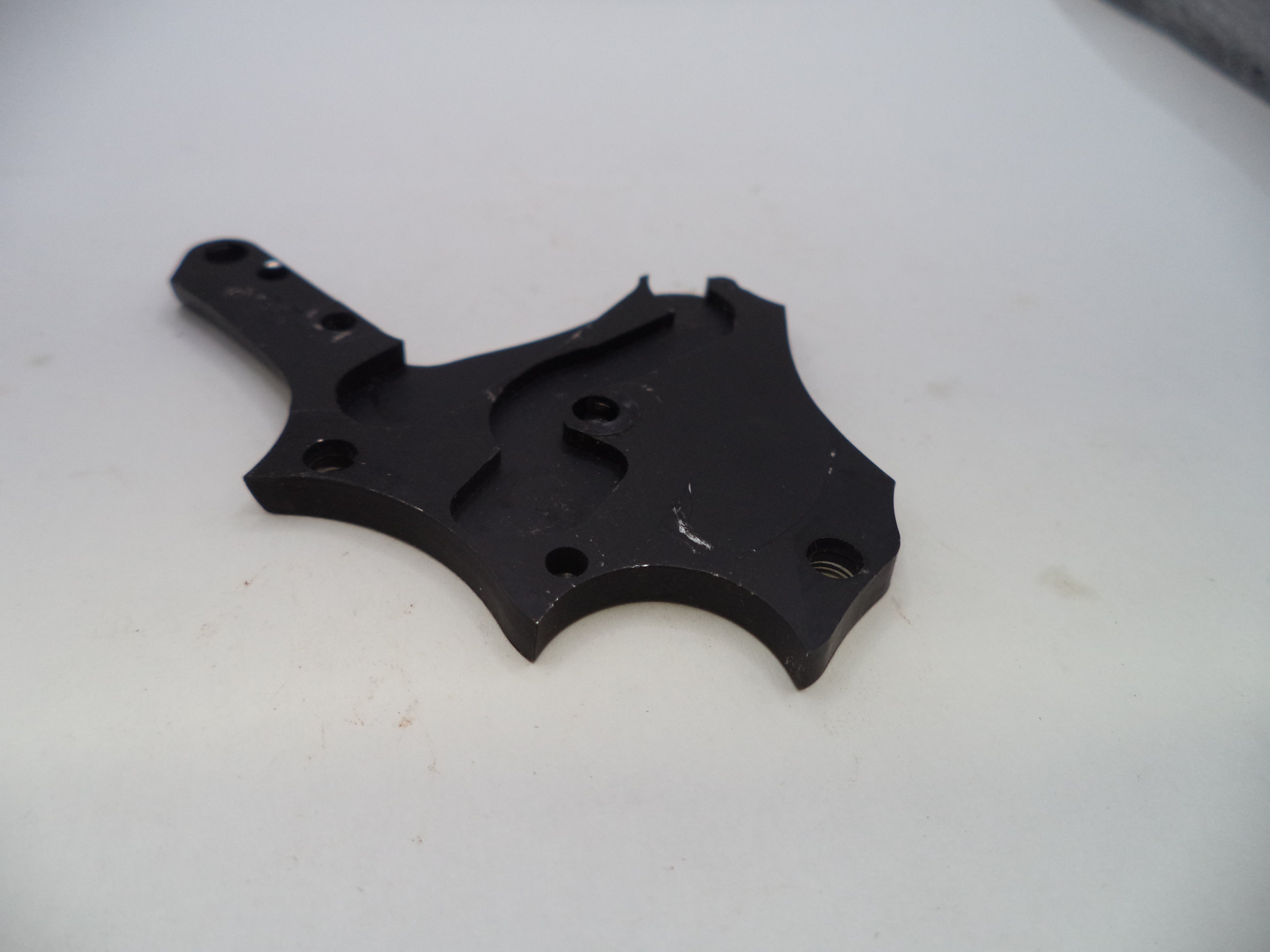 A black metal mechanical bracket or hardware piece with pre-drilled holes, photographed on a plain off-white surface.