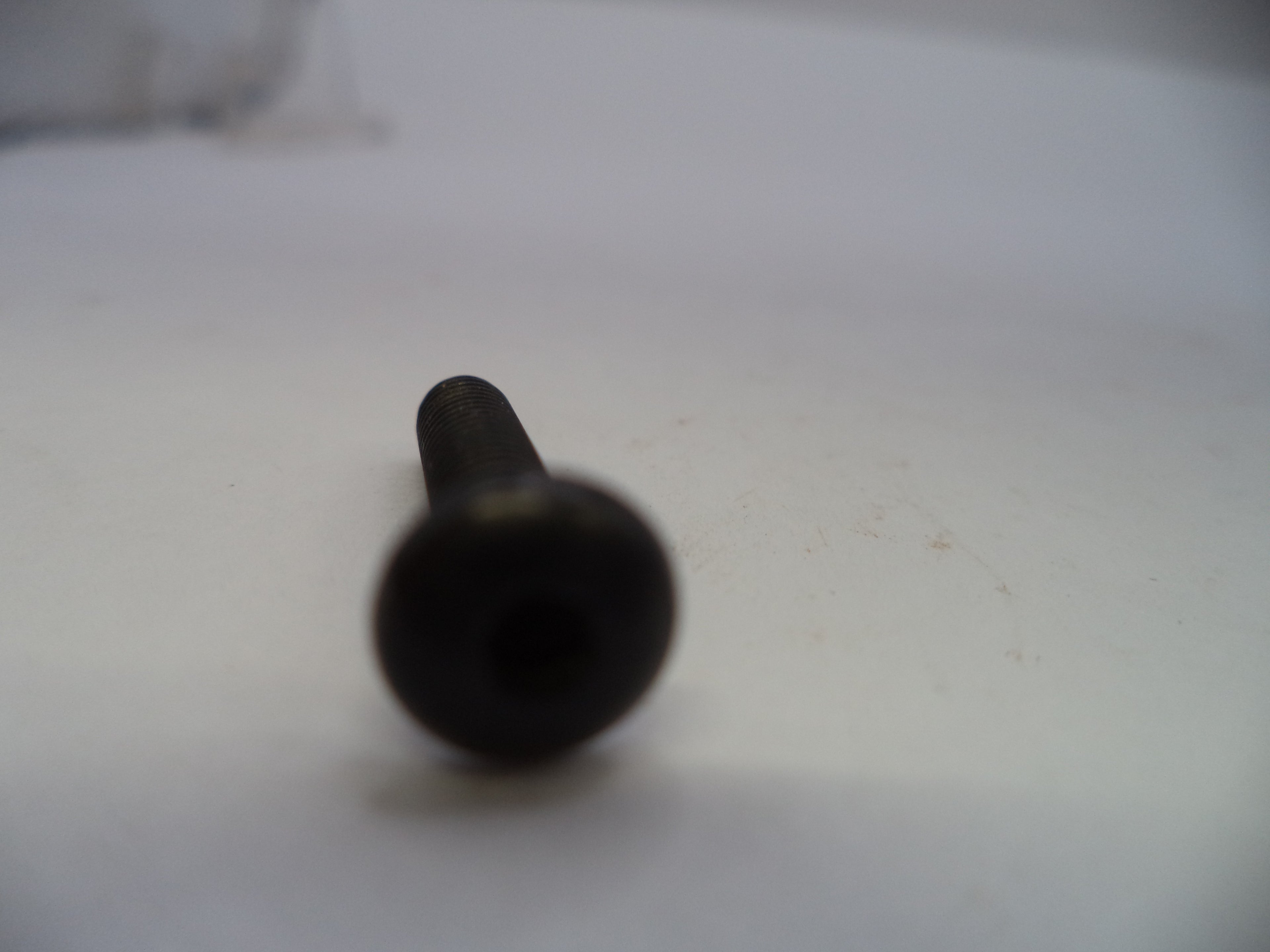 A single black metal screw or bolt photographed on a plain light background, with the round head in sharp focus and the shaft blurred due to shallow depth of field.