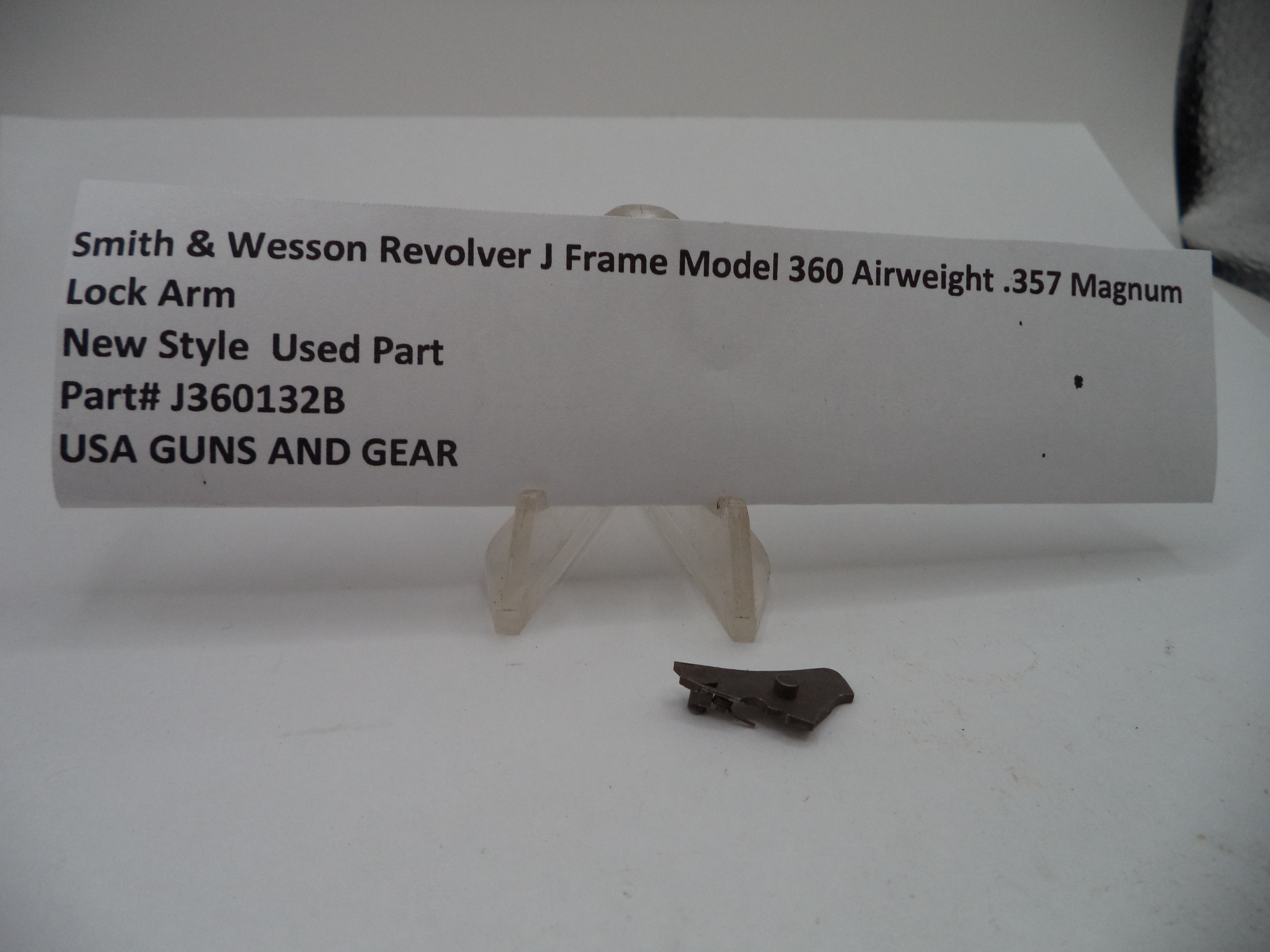 A small used metal lock arm part displayed on a clear plastic stand in front of a white paper label card. The label reads 'Smith & Wesson Revolver J Frame Model 360 Airweight .