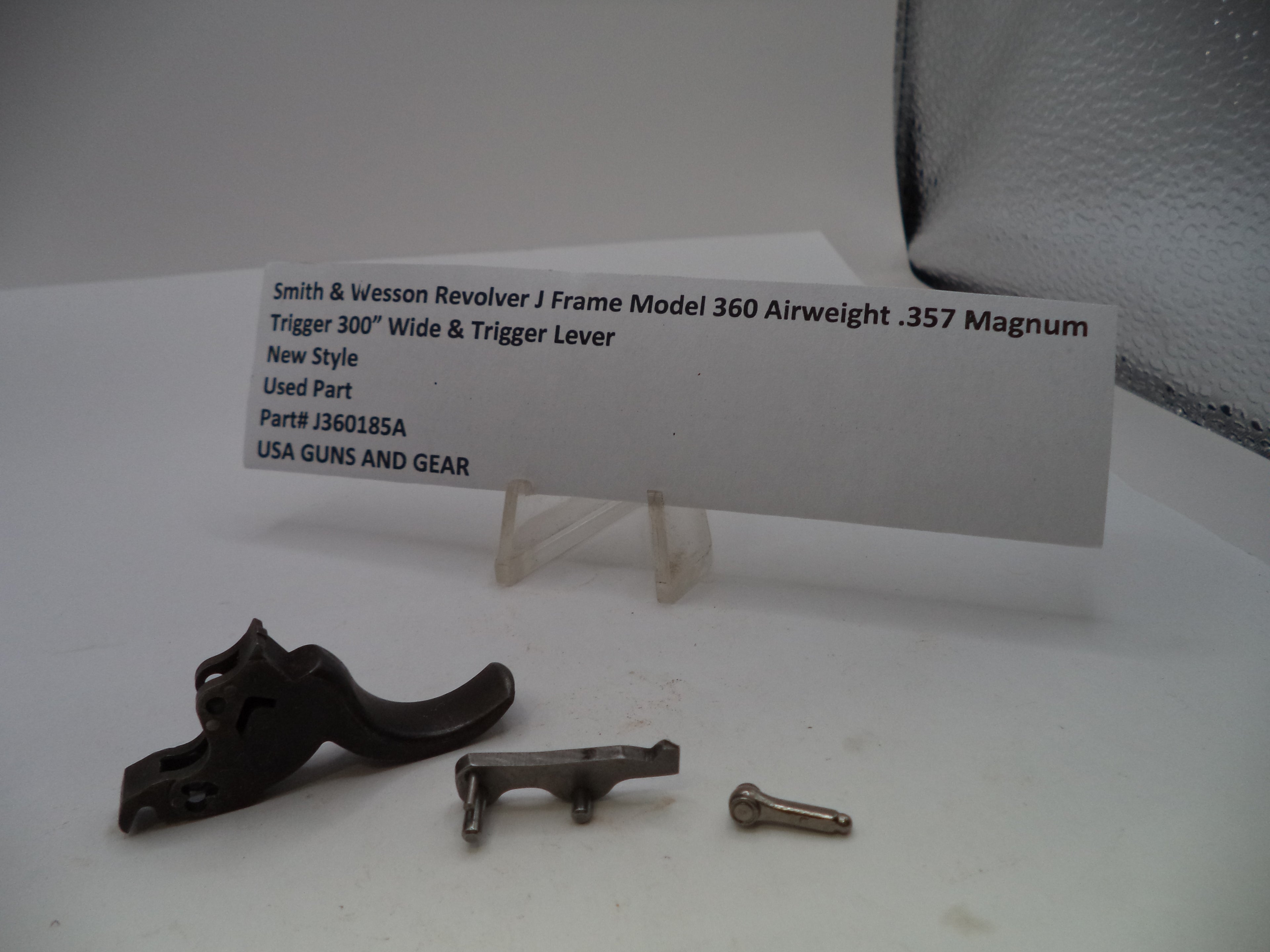 A used Smith & Wesson revolver J frame trigger lever set for a .357 Magnum, including the black trigger lever, a silver metal piece, a small screw, and a white product information card on a clear stand.