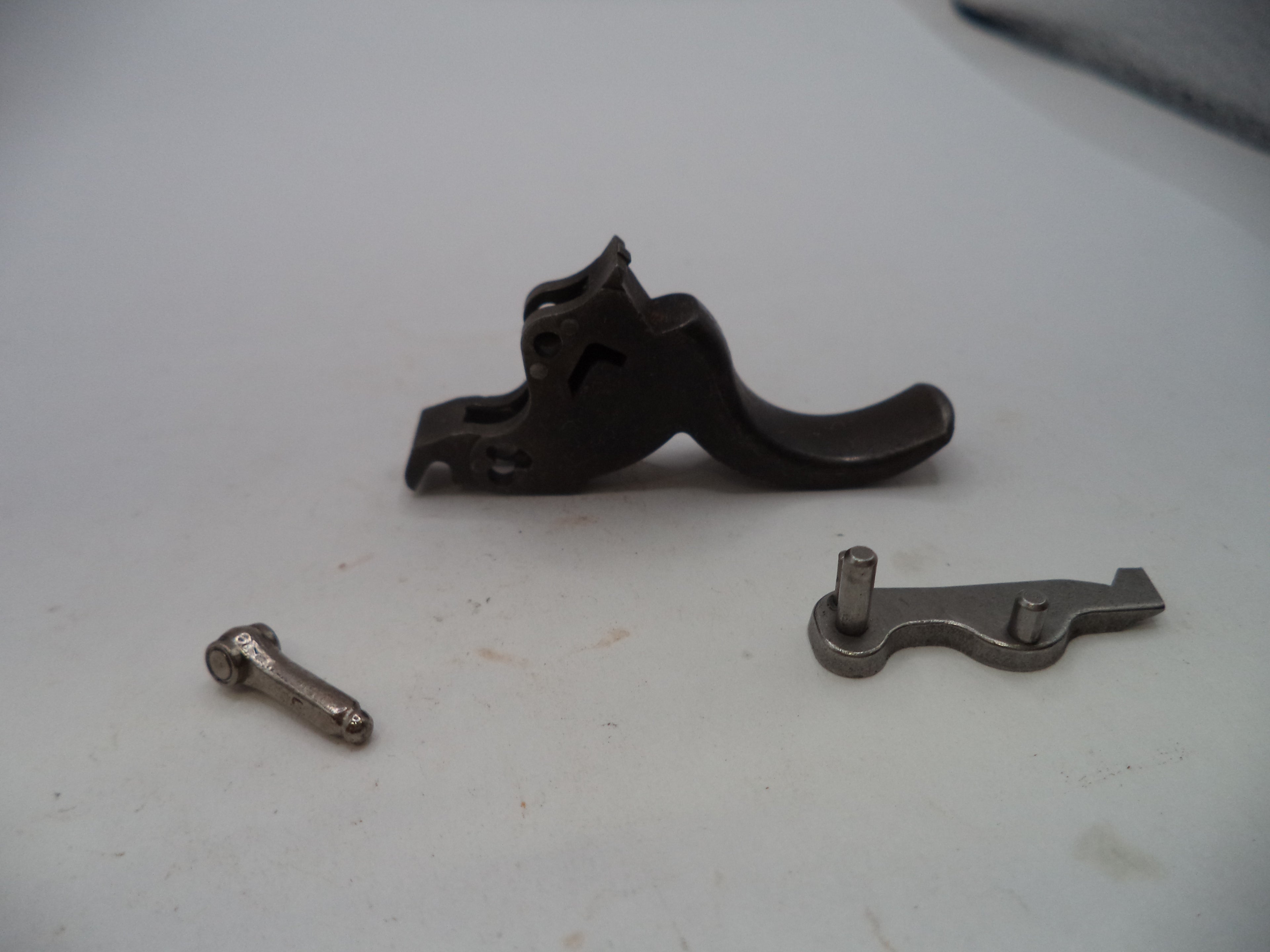 A close-up of a disassembled firearm trigger assembly, showing a dark metal trigger assembly, a separate silver-toned spring, and a small silver-toned screw on a light gray surface.