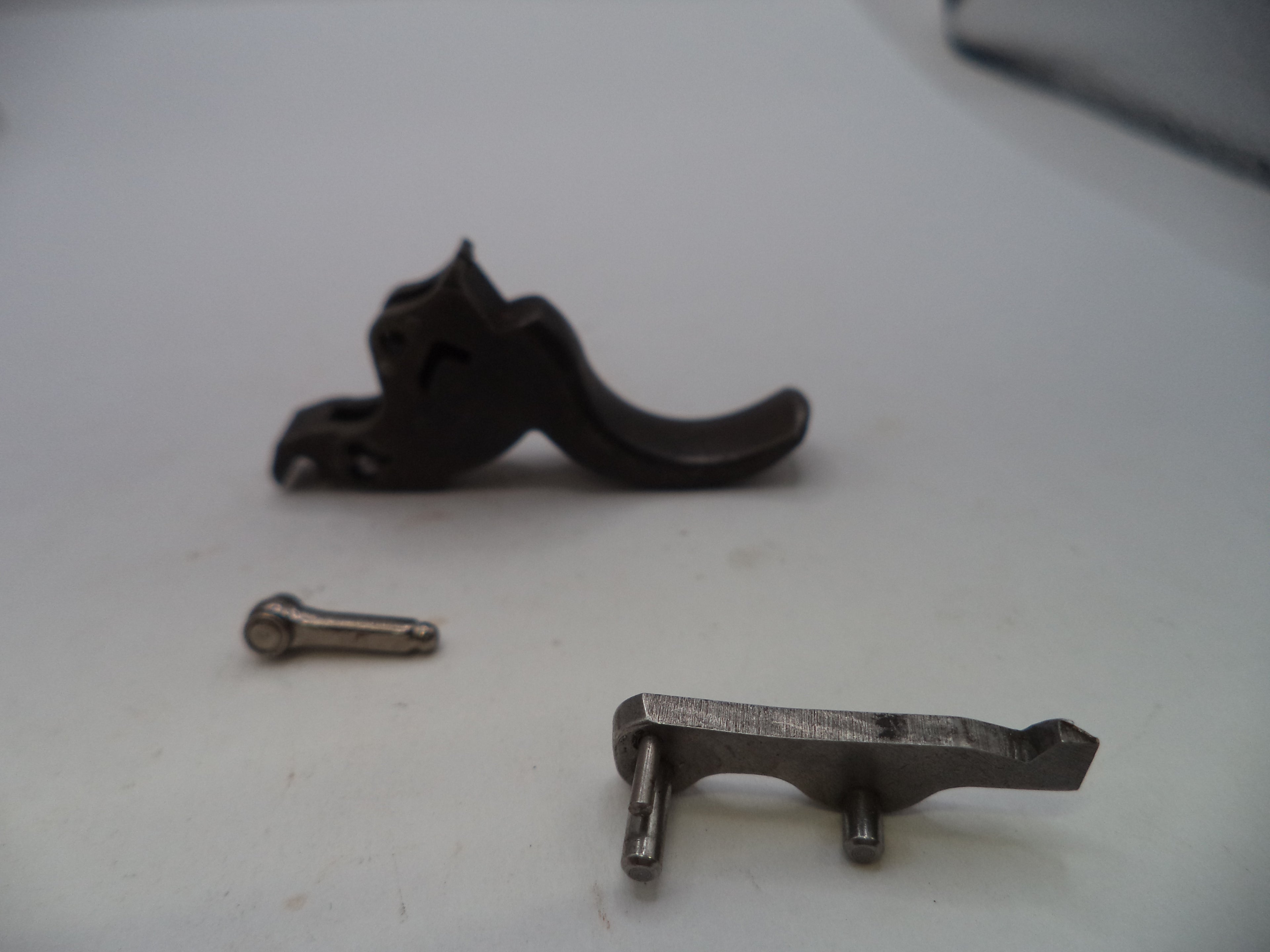 A set of firearm replacement parts, including a dark metal trigger assembly, a separate silver-toned pin, and a matching silver-toned lever, displayed on a plain white surface.