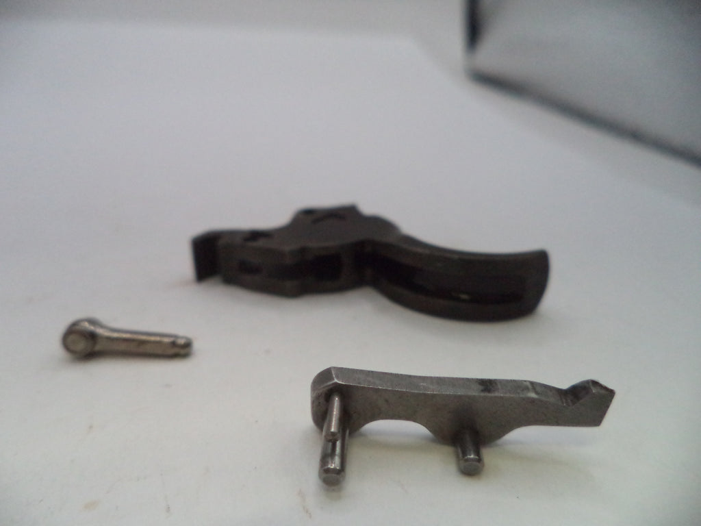 A close-up shot of a disassembled metal trigger assembly, showing a dark metal trigger component, a separate metal spring, and a metal pin on a white surface.