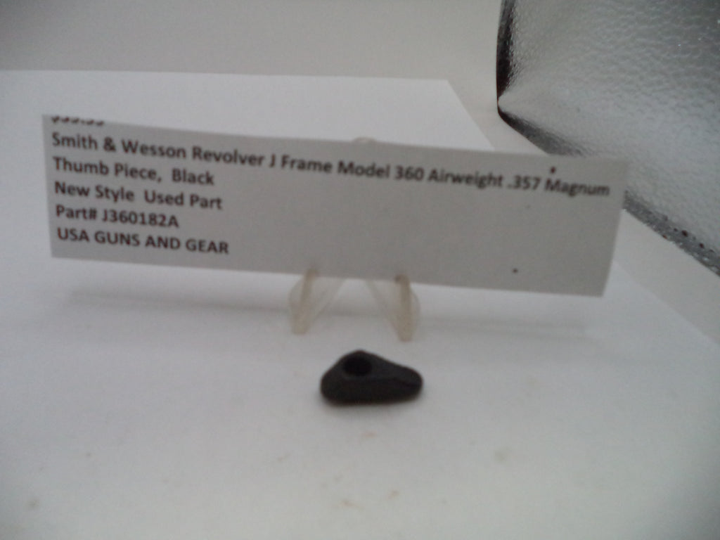 A small black plastic thumb piece for a Smith & Wesson revolver, displayed on a clear plastic stand next to a white paper label with product details.