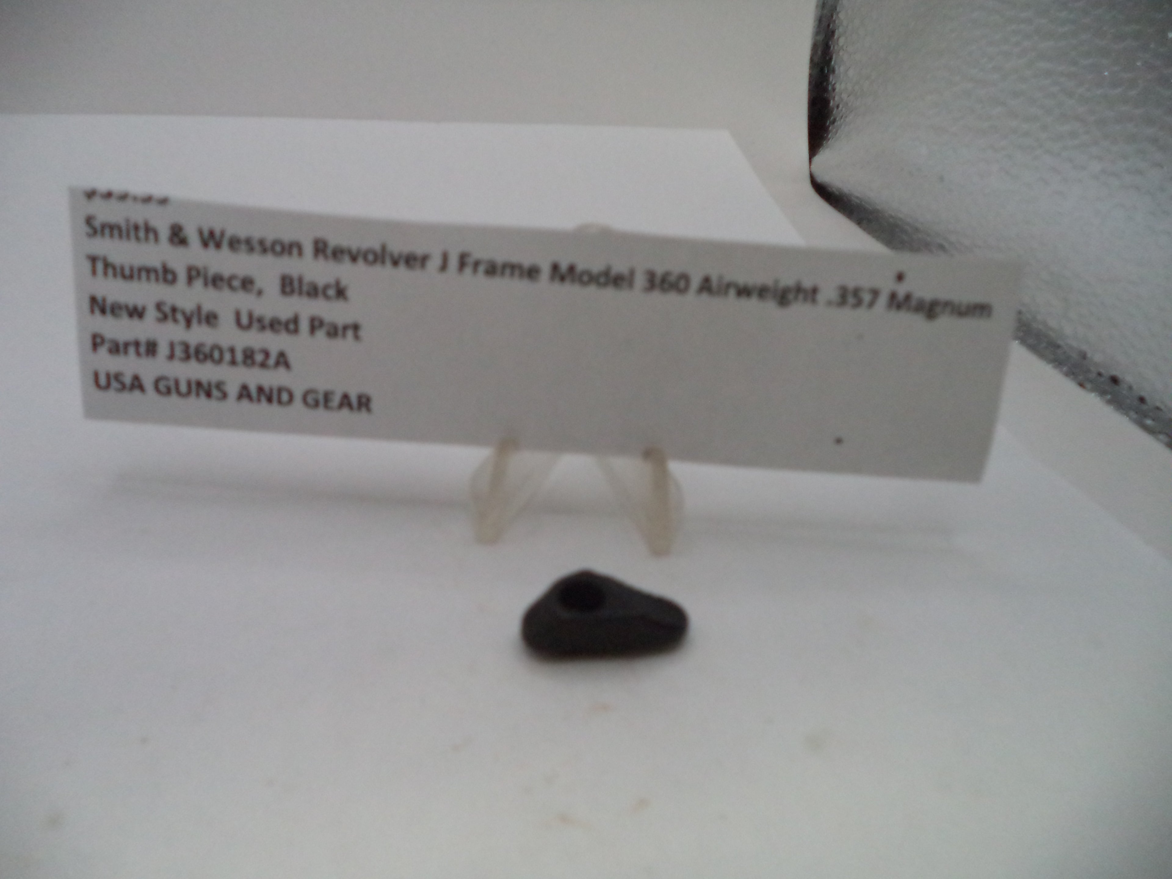 A small black plastic thumb piece for a Smith & Wesson revolver, displayed on a clear plastic stand next to a white paper label with product details.