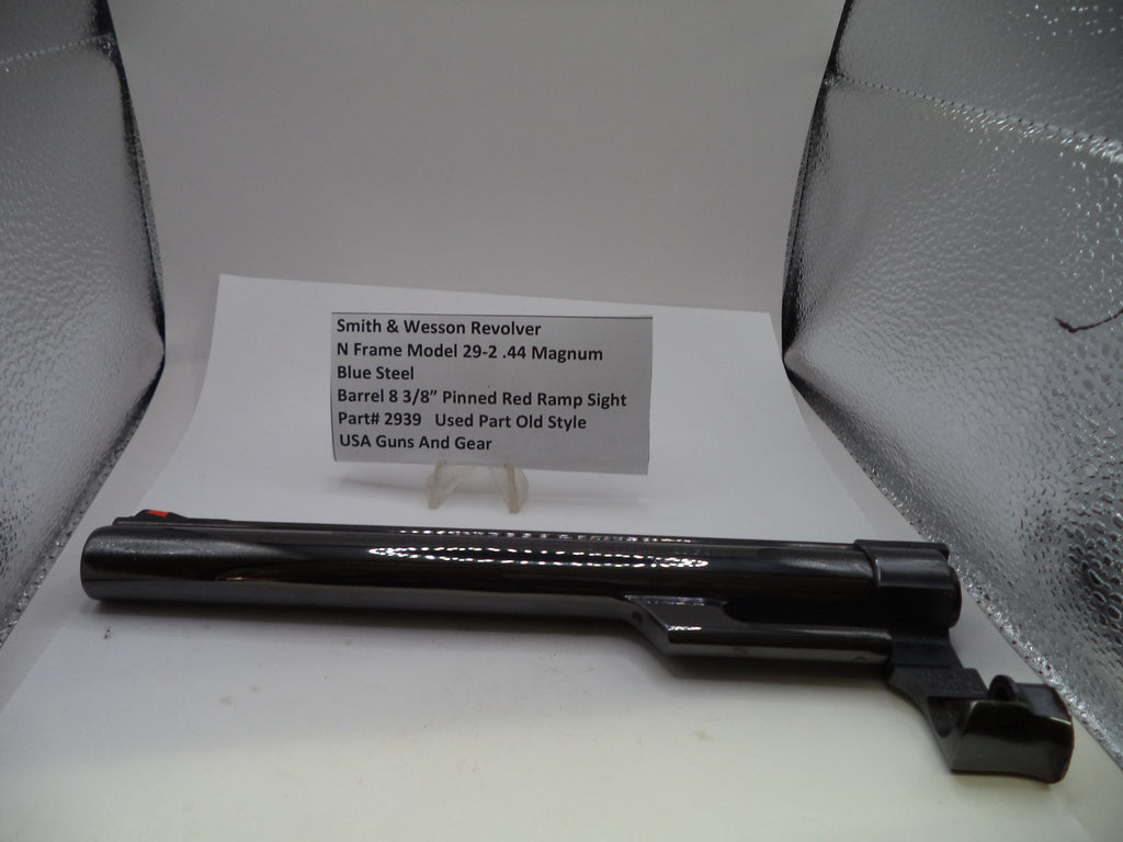 A black Smith & Wesson revolver receiver or barrel component displayed on a white surface with a printed parts information card behind it, set against a silver reflective backdrop.