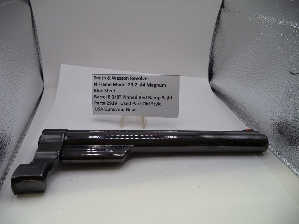 A black Smith & Wesson revolver barrel and frame component displayed on a white surface with a printed part information card behind it, set against a silver textured background.