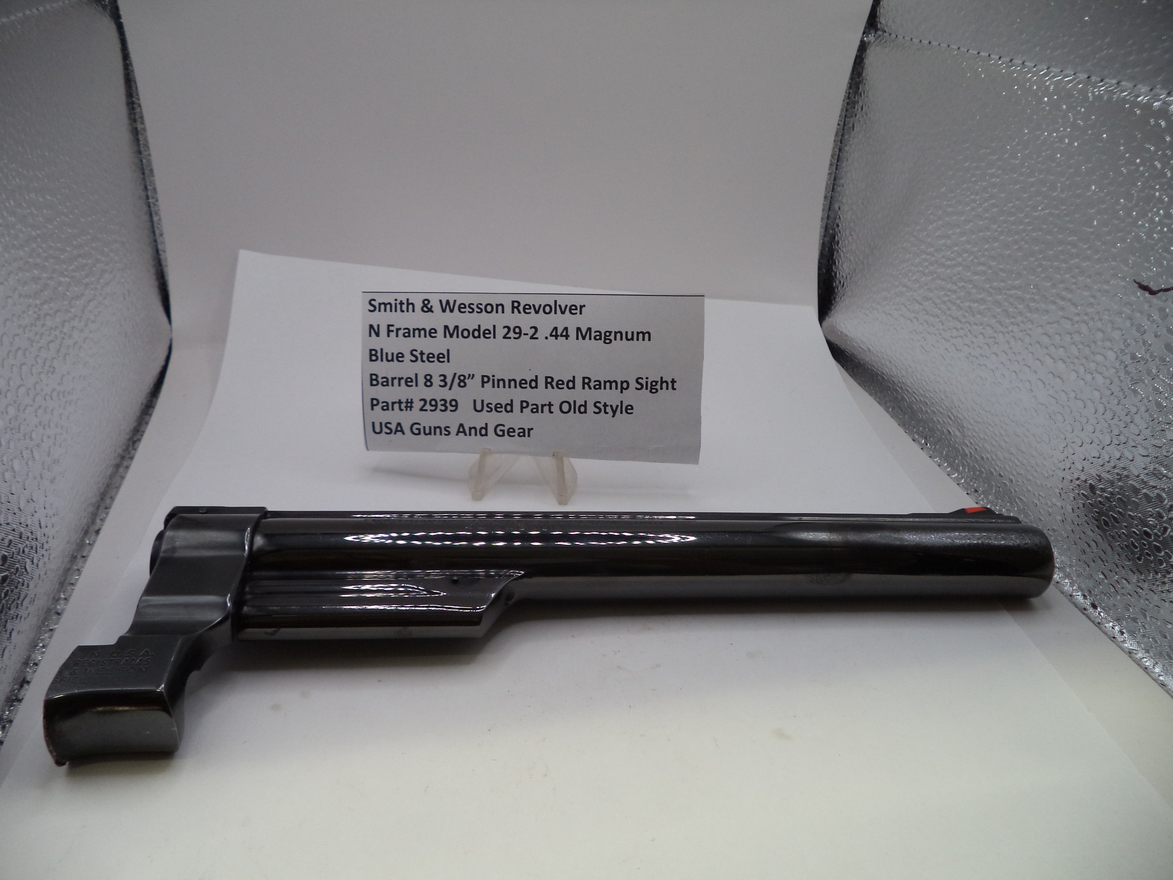A black Smith & Wesson revolver barrel and frame component displayed on a white surface with a printed part information card behind it, set against a silver textured background.