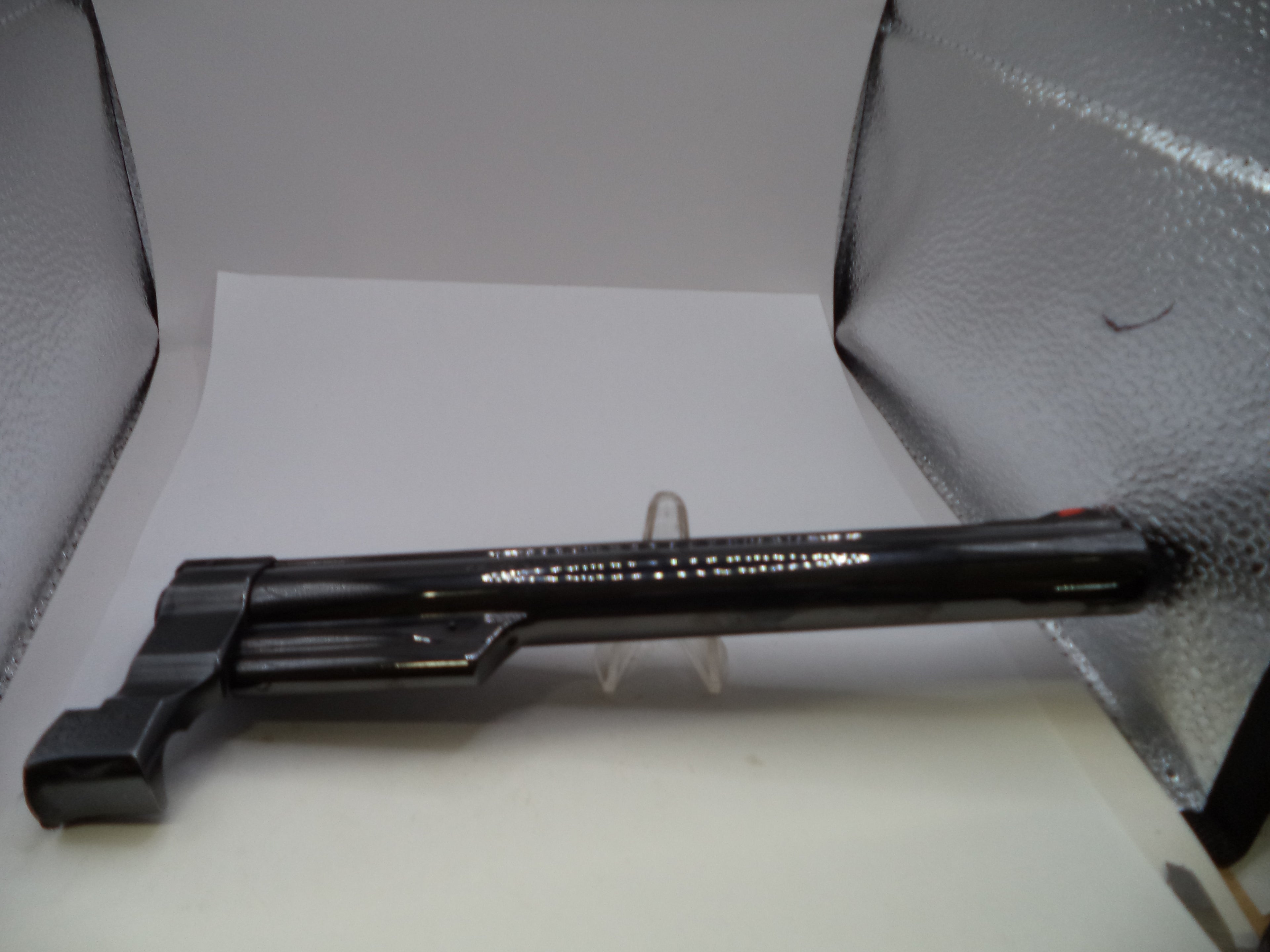 A black plastic toy gun prop with white printed text, displayed on a small clear stand inside a lightbox with silver reflective walls.