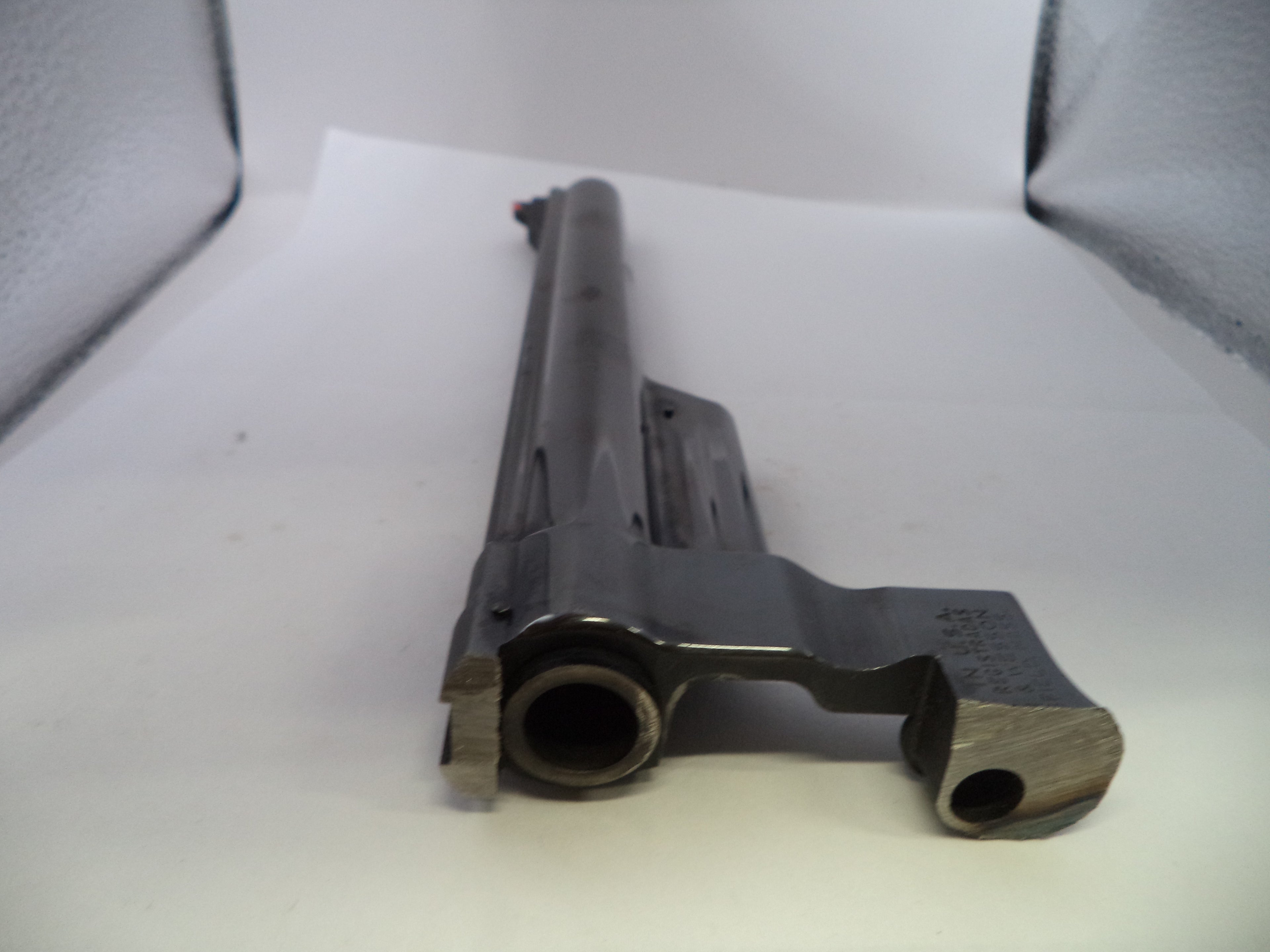 A close-up of a dark metallic gun receiver and barrel section photographed on a white surface with a textured gray backdrop.