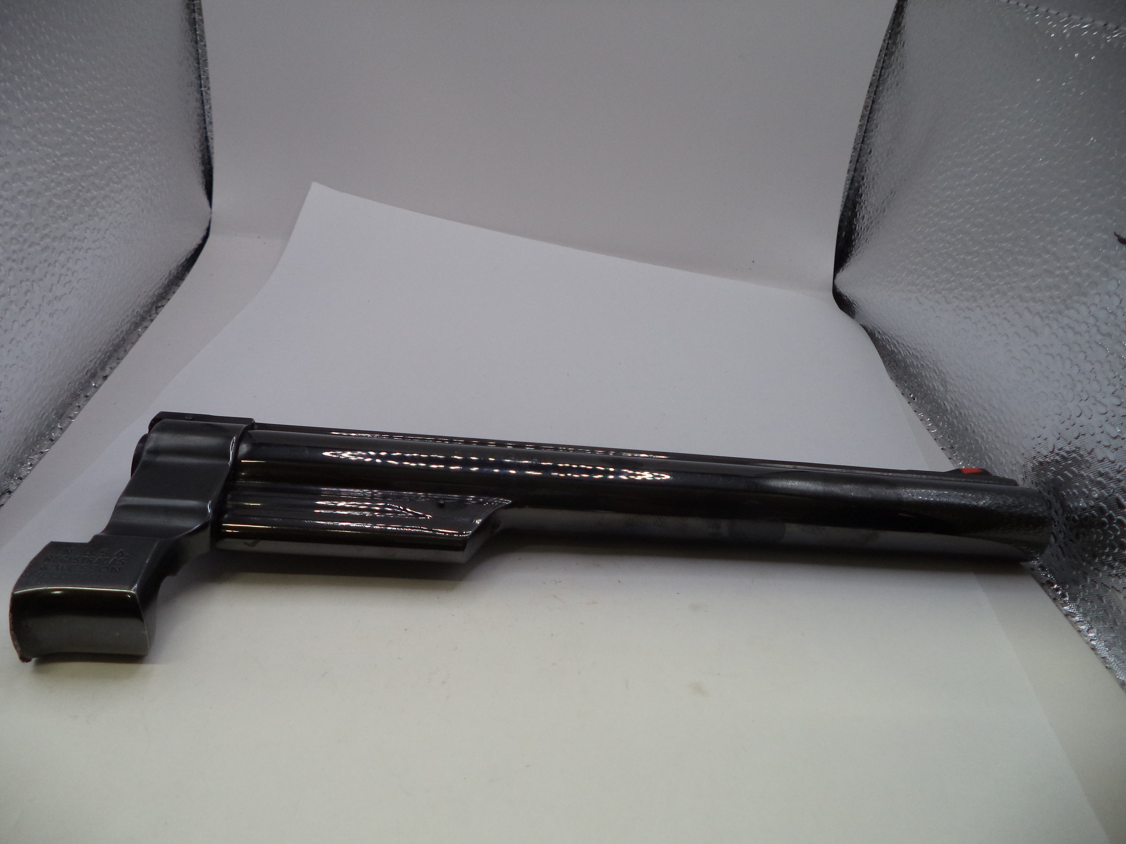 A black plastic toy gun or prop blaster with a textured grip, lying on a white sheet of paper inside a silver lightbox.