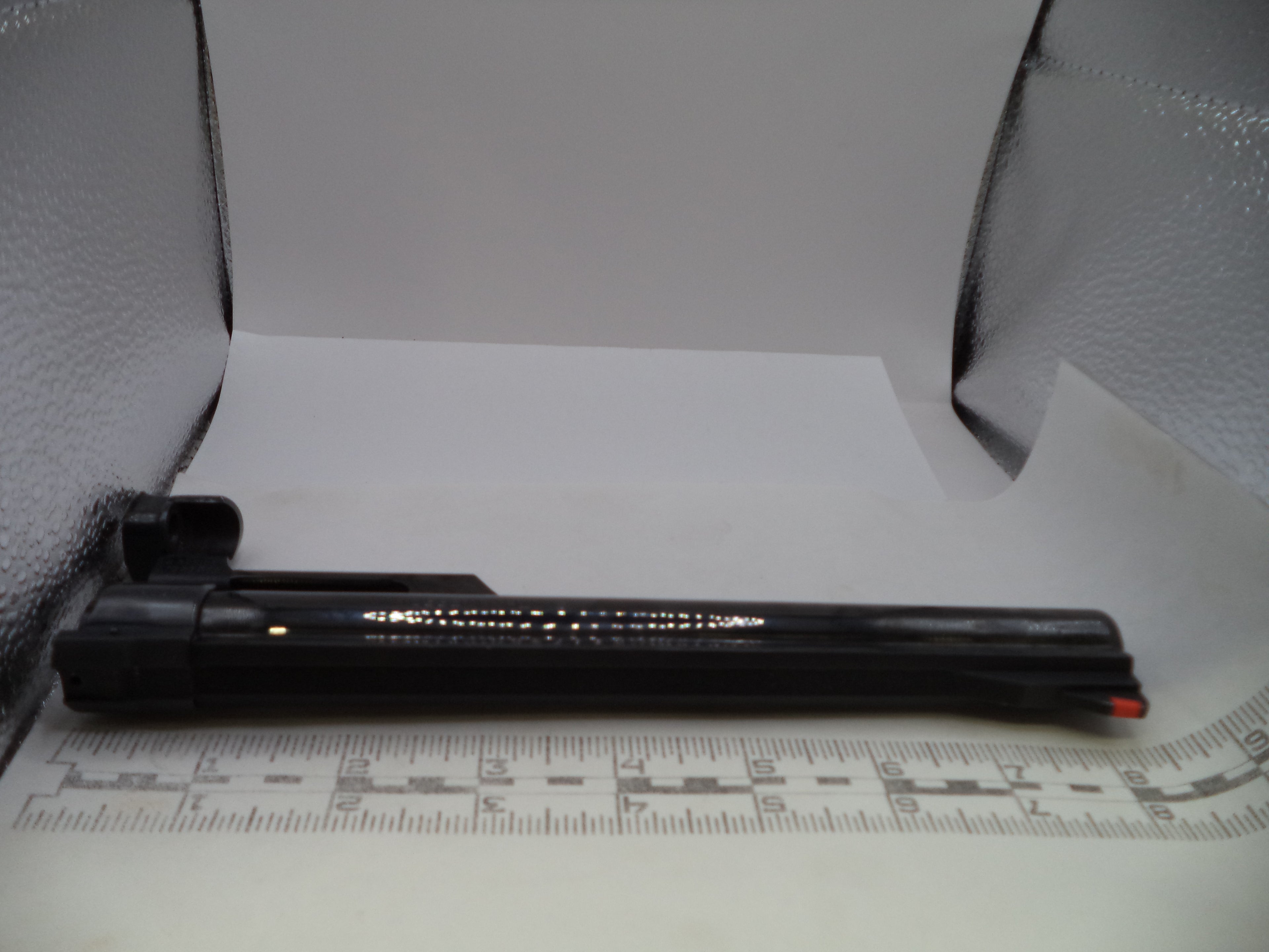 A black rifle scope mount or accessory bar with gold lettering, shown on a white background with a measuring ruler in front and textured silver walls behind.