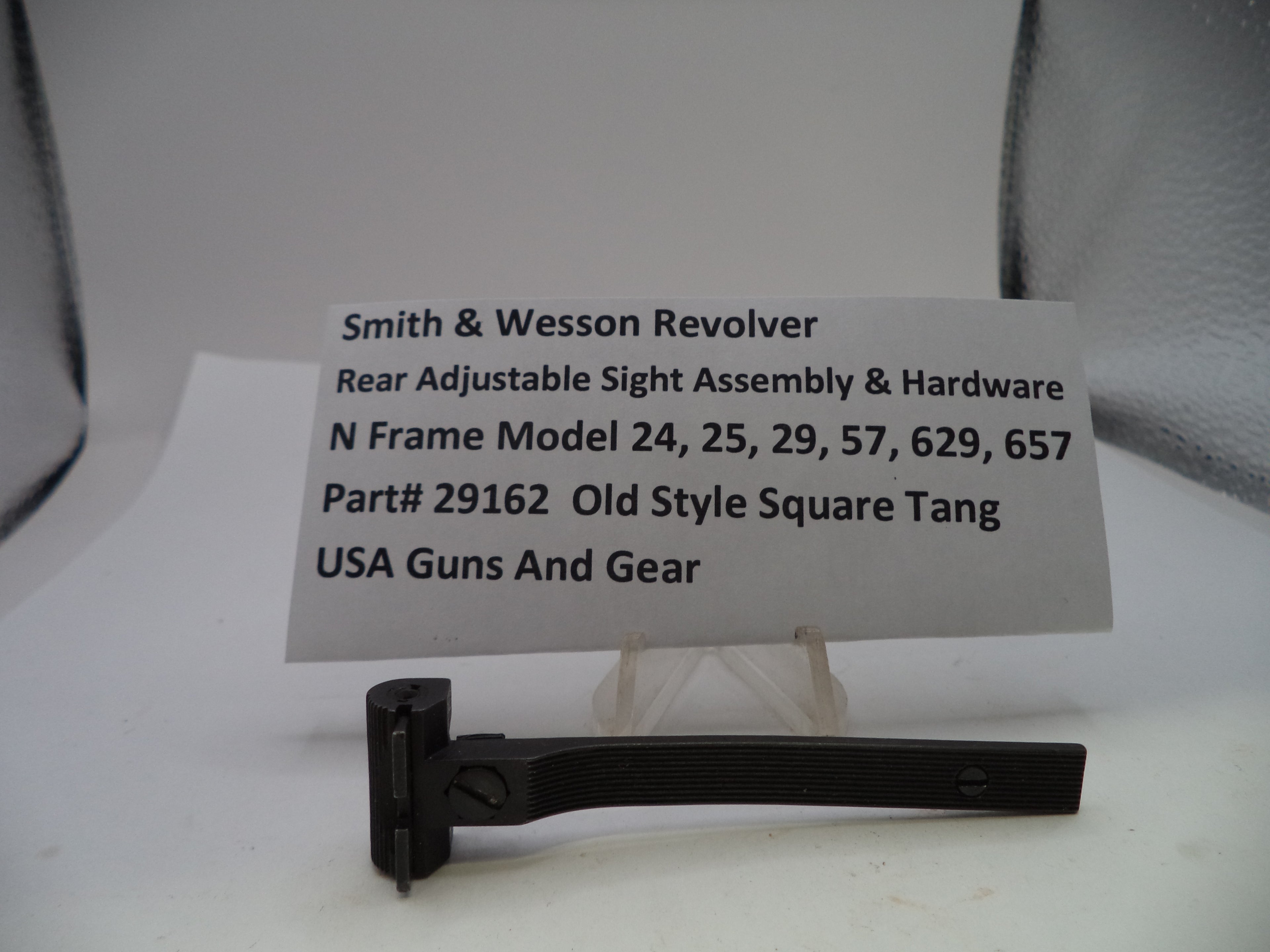 A black metal Smith & Wesson revolver rear adjustable sight assembly and hardware displayed on a small clear stand, with a white label card behind it showing product details.