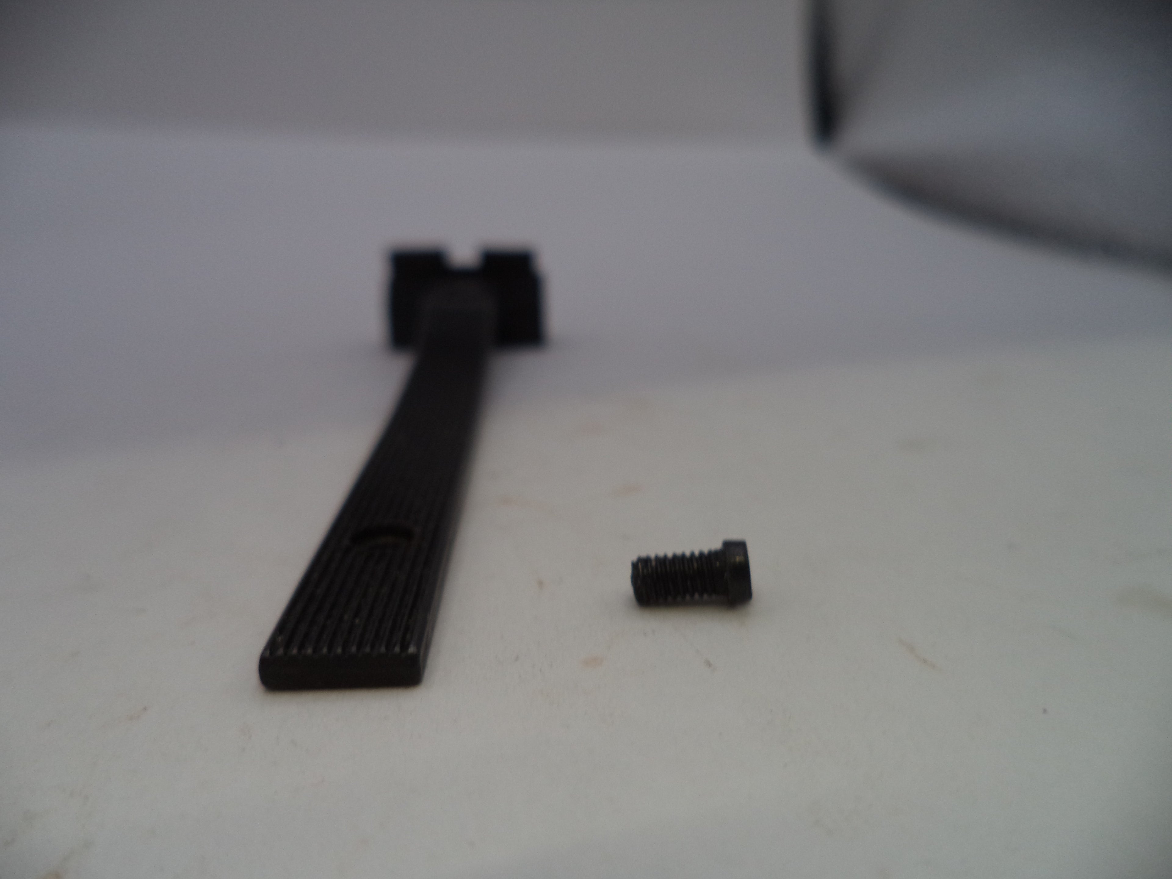 A black metal mounting bar with a ribbed textured surface and a small black screw placed beside it on a light gray surface.