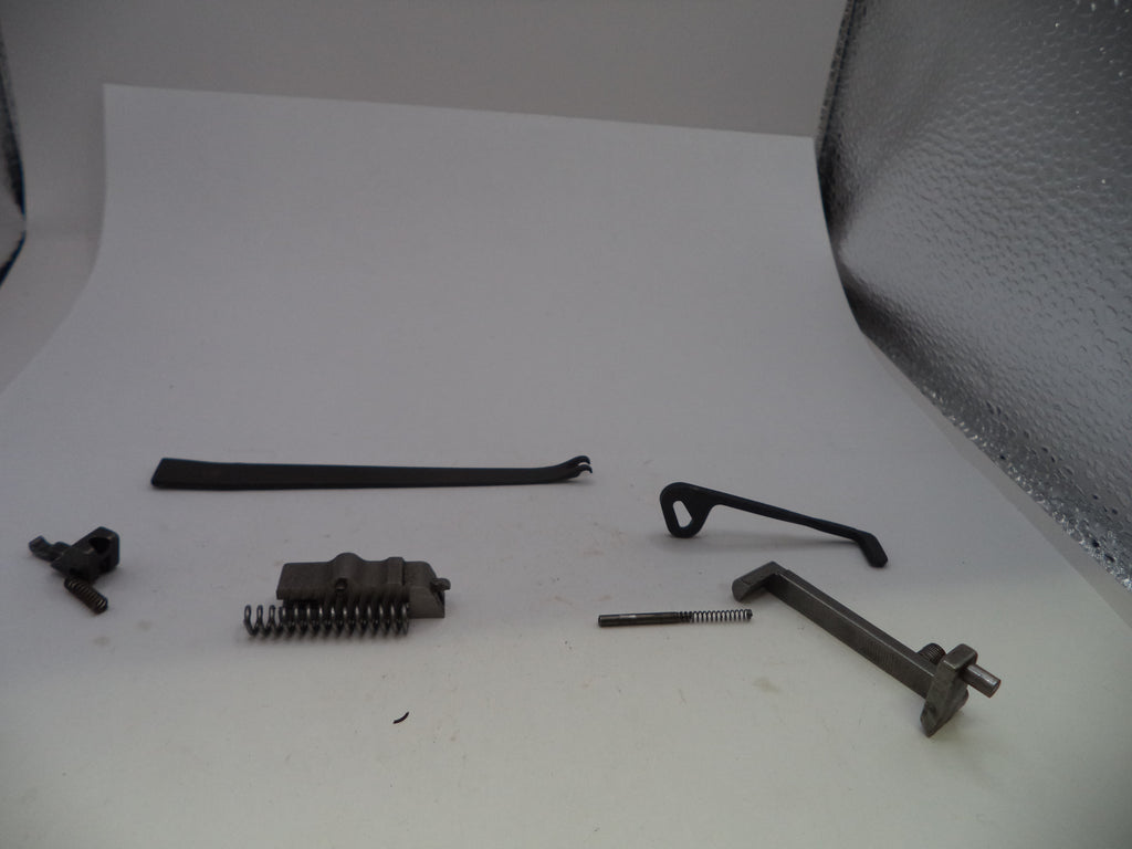 A set of small dark metal mechanical parts and springs arranged on a white studio surface, with a textured silver backdrop visible behind them.