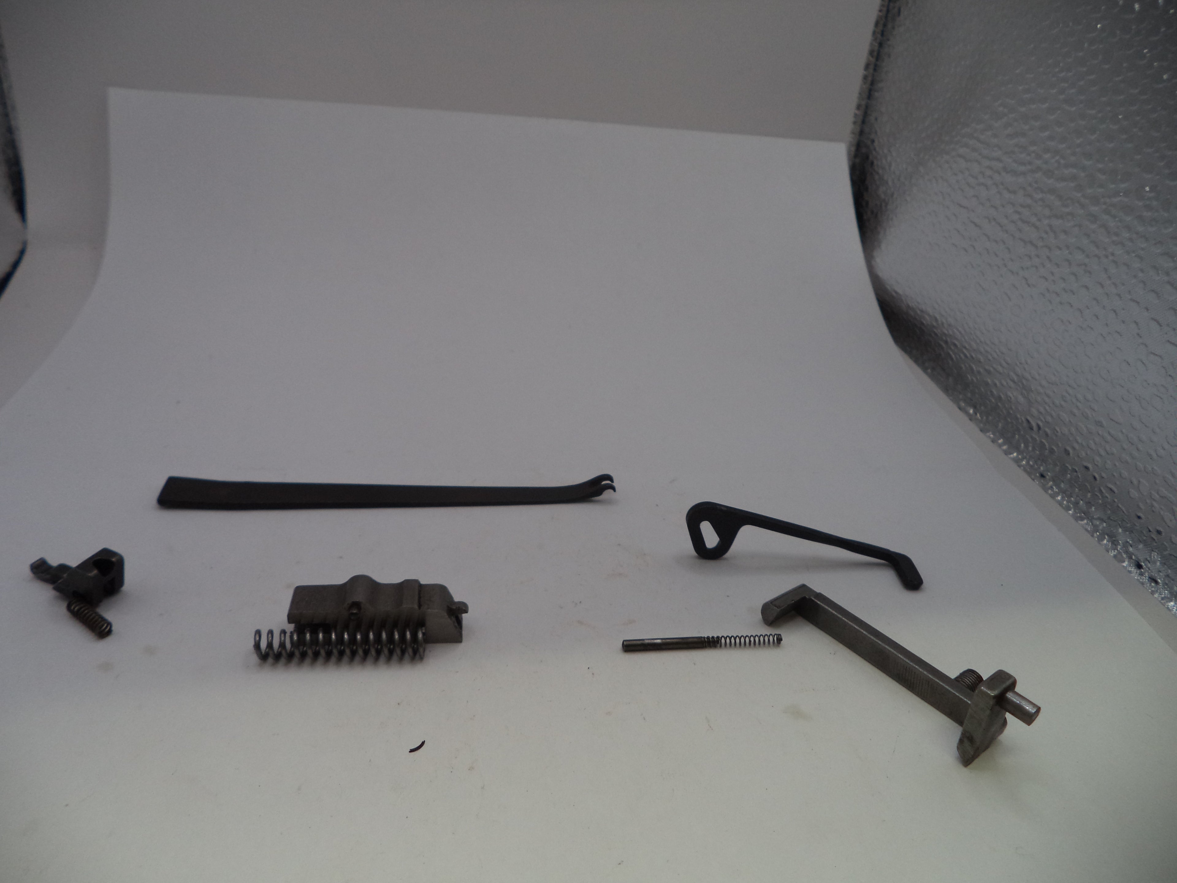 A set of small dark metal mechanical parts and springs arranged on a white studio surface, with a textured silver backdrop visible behind them.