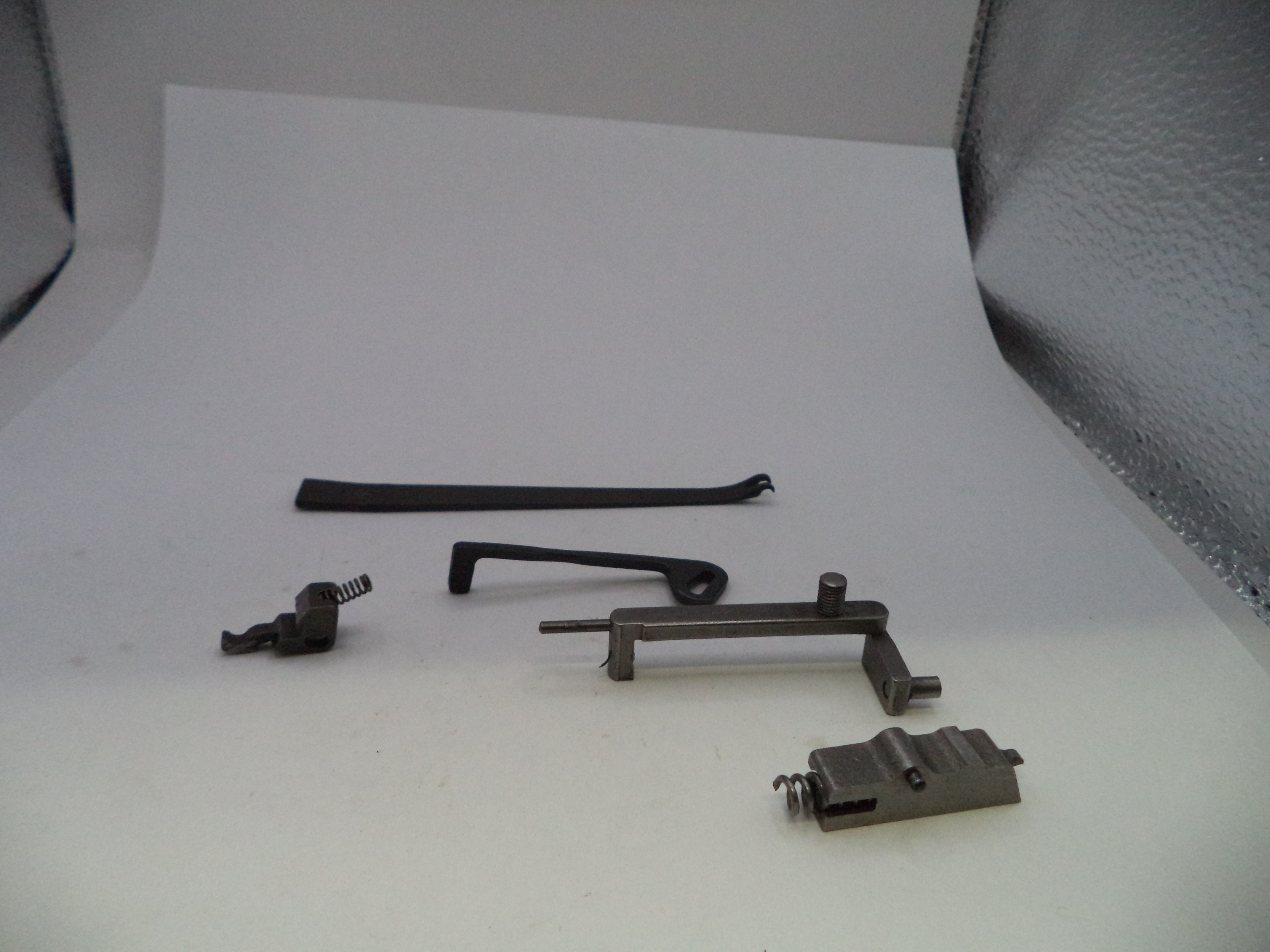 A set of small metal and black hardware components, including a long black strip, a dark metal bracket, a small black spring piece, and a gray metal latch or clip, arranged on a white studio background.