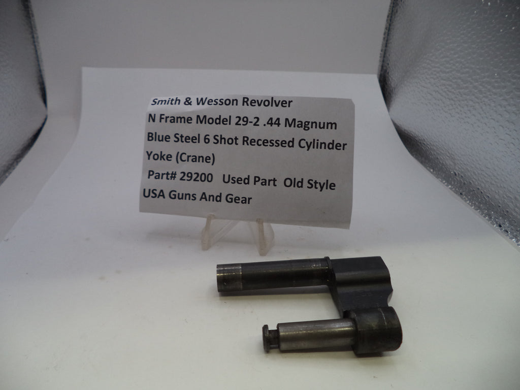 A used Smith & Wesson revolver cylinder yoke part, made of blue steel, displayed on a small clear stand next to a paper label card with product details.