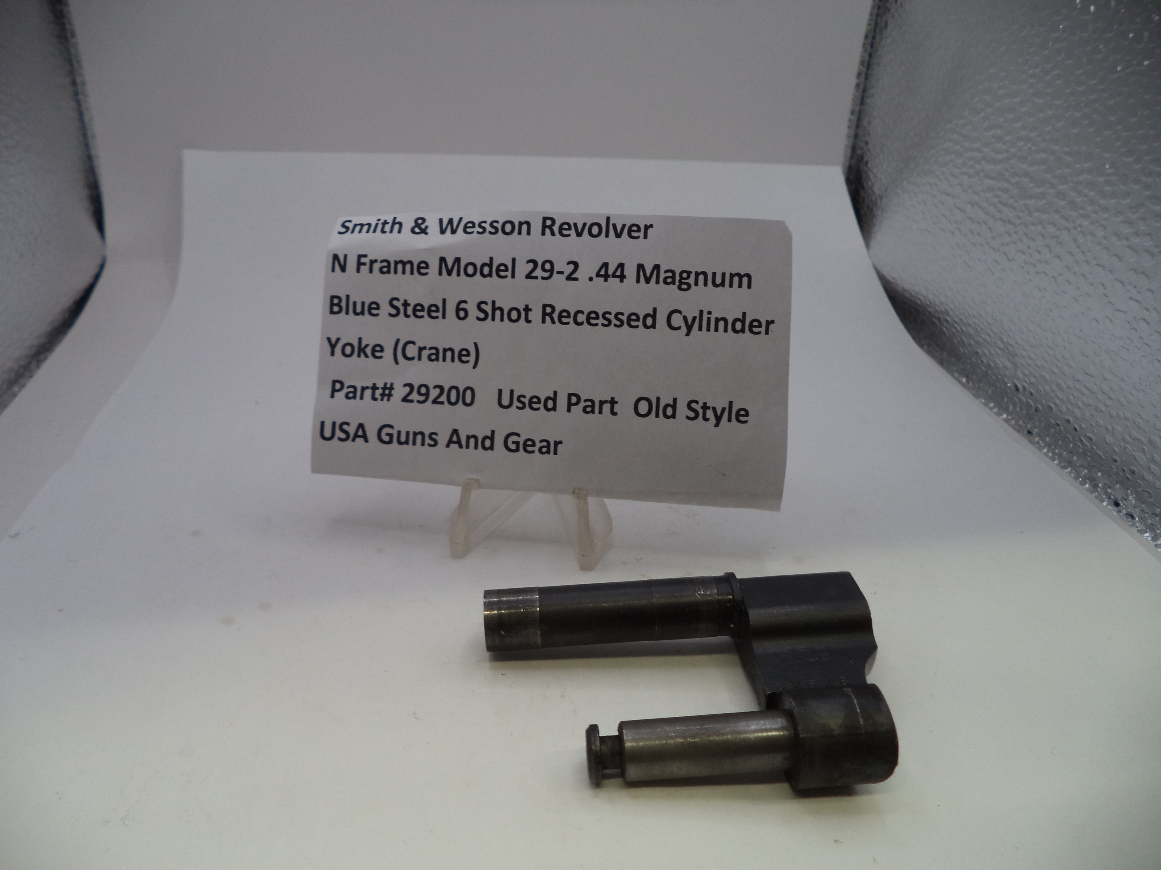 A used Smith & Wesson revolver cylinder yoke part, made of blue steel, displayed on a small clear stand next to a paper label card with product details.