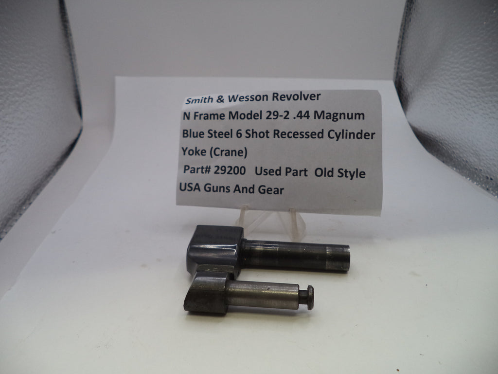 A used Smith & Wesson revolver cylinder yoke part displayed on a white surface with a handwritten label card behind it. The part consists of two dark blue steel components with machined silver metal ends.